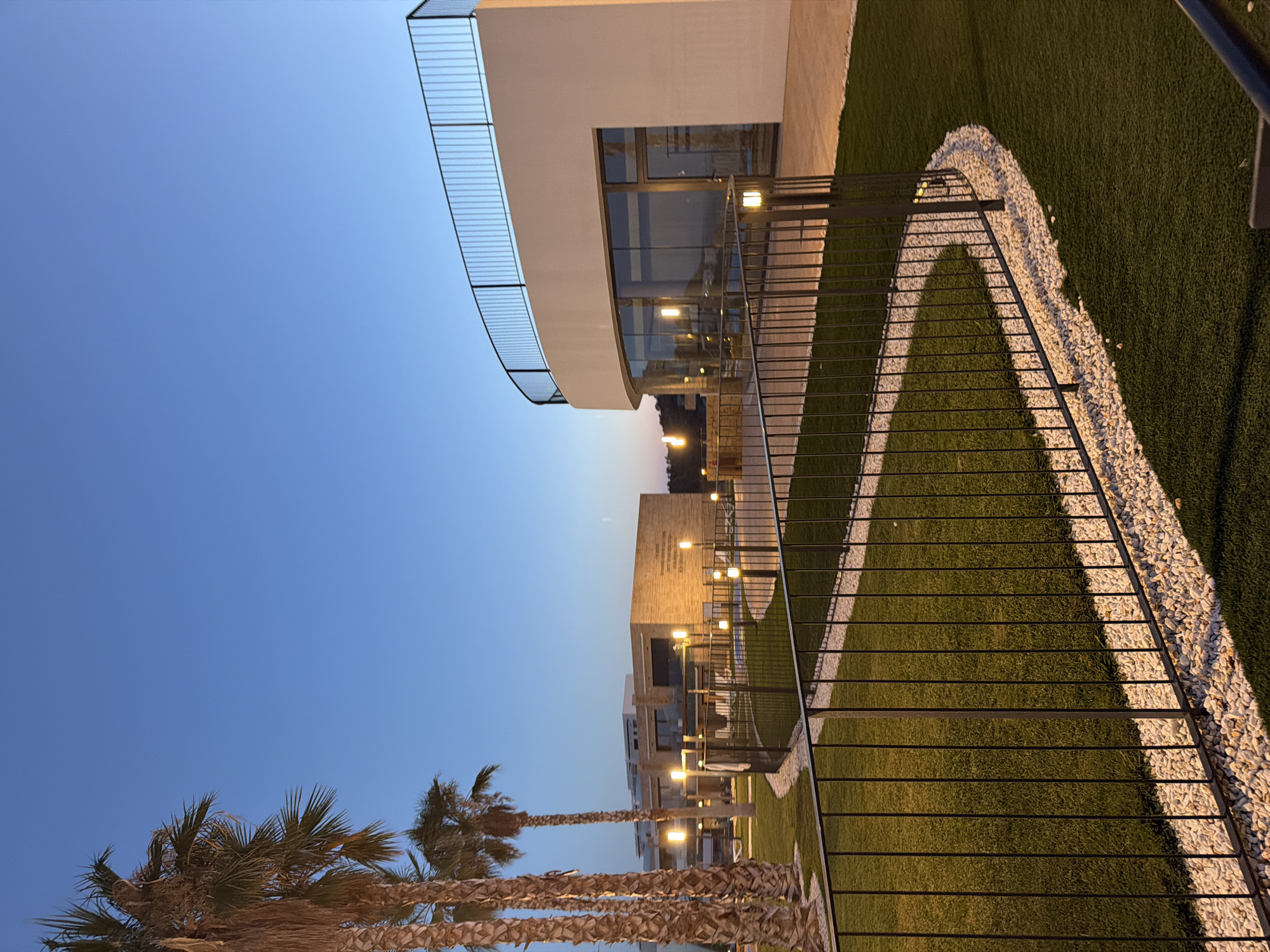 The image captures a modern architectural complex in Finestrat, Spain, at dusk. Three palm trees stand tall on the left, their fronds silhouetted against a clear blue sky. To the right, a sleek, curved building features large glass windows and a black railing on its upper level. In the background, another building with a brick facade is illuminated by outdoor lighting, as is a pathway lined with small white stones and artificial turf. A black metal fence encloses a section of the manicured lawn. The scene is peaceful, with no people visible.