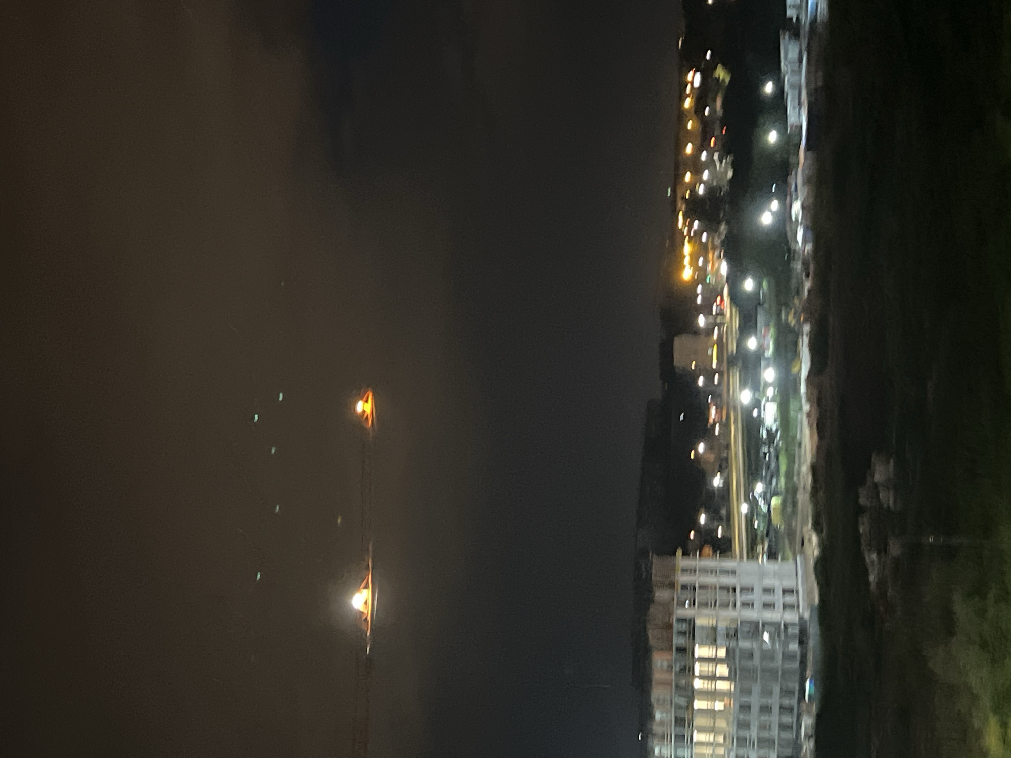 The image depicts a night scene in Yekaterinburg, Russia, likely from a high vantage point, possibly a window. The dominant elements are a dark, somewhat cloudy sky and a city illuminated by artificial lights. In the foreground, two glowing lights, possibly street lamps or decorative lights, are visible against the dark sky, with one being significantly brighter and closer. These lights appear to be suspended over a body of water, with subtle reflections visible.
To the right of the frame, a cityscape unfolds. A multi-story building, appearing to be under construction or recently built, is prominent with many windows lit from within, suggesting occupancy. Further into the distance, a line of lights runs horizontally, indicative of roads or another row of buildings. These distant lights are a mix of warm yellow and cooler white, creating a blurred effect due to the nighttime and distance. There is also a suggestion of greenery or trees to the far right of the city lights.
The overall atmosphere is tranquil and urban. The time of day is clearly night, and the weather appears calm, with the lights reflecting subtly on an unseen surface. No people are clearly discernible, and the focus is on the illuminated urban landscape against the dark sky. No text is visible in the image.