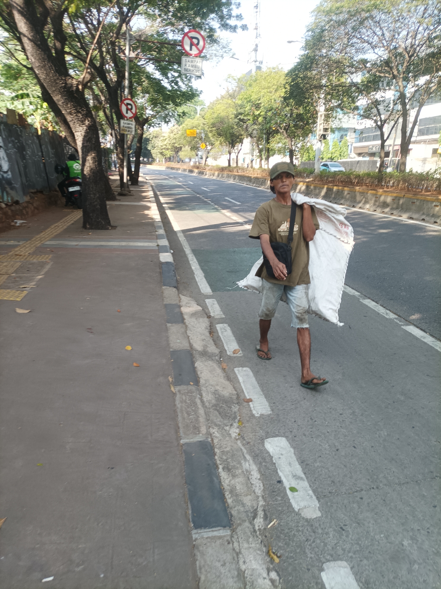In South Jakarta, Indonesia, a man walks along the side of a road, carrying a large white sack over his shoulder. He is wearing a tan t-shirt, light-colored shorts, a cap, and flip-flops. A black bag is slung across his chest. The man is walking on a paved sidewalk next to a busy road with multiple lanes of traffic. Trees line the sidewalk, and buildings are visible in the background. Two "No Parking" signs are posted on poles along the roadside. The scene appears to be on a sunny day, with shadows cast by the trees.
