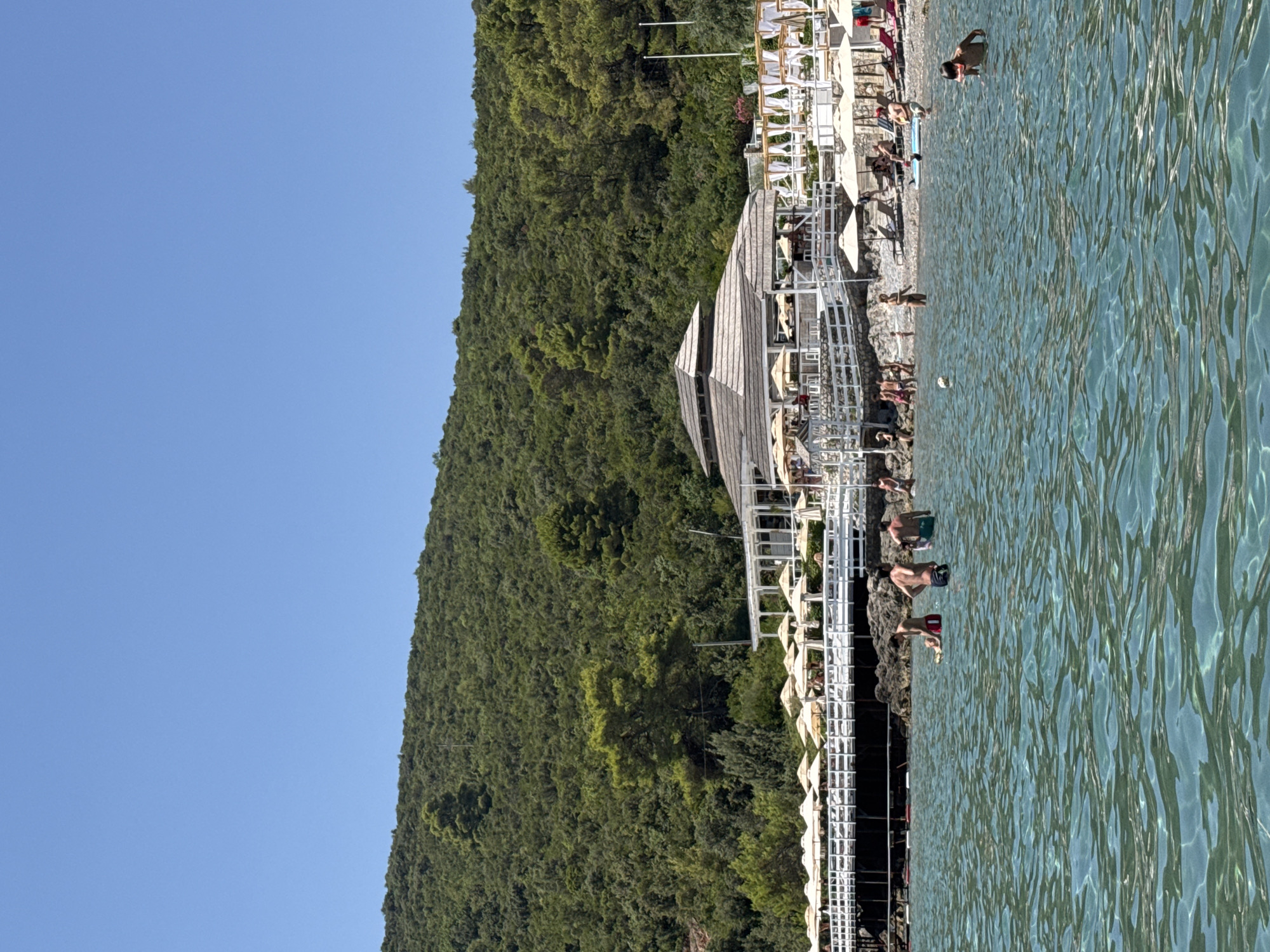 A bright, sunny day is depicted at a beach resort located in Radovići, Montenegro. The foreground features clear, turquoise water where several people are swimming and wading. In the middle ground, a pebble beach is visible, lined with numerous white lounge chairs and beige umbrellas, suggesting a resort or private club. A white wooden structure with a thatched roof and open-air seating area stands prominently behind the beach. Further back, a densely forested green hillside rises, with a clear blue sky overhead. People are scattered across the beach, some sunbathing, others socializing, and a few are in the water. A white railing delineates the edge of the beach area. The overall scene captures a lively, recreational atmosphere at a coastal resort.