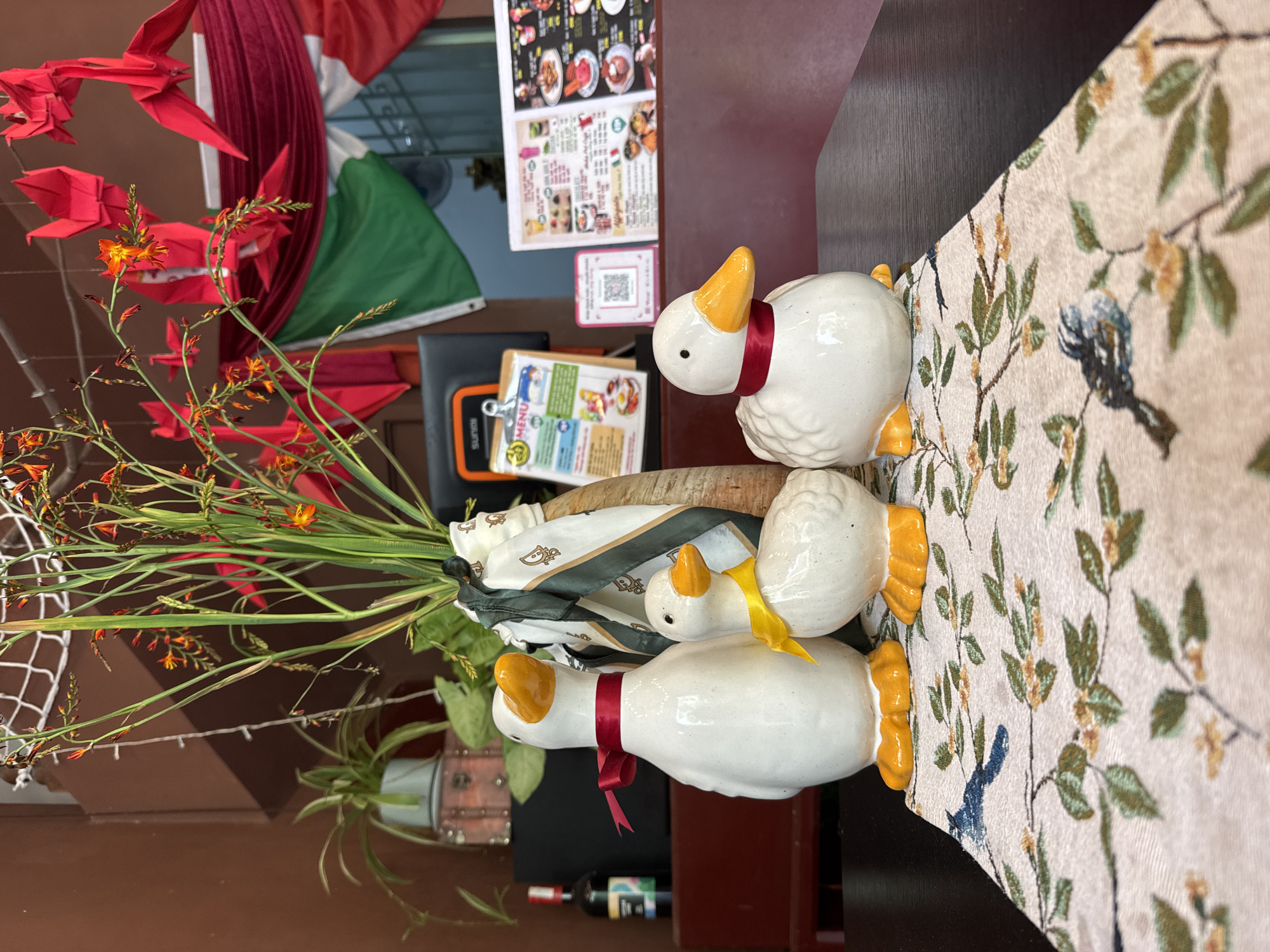 The image shows a table setting in what appears to be a restaurant or dining area, potentially in Nha Trang Ward, Vietnam. Three white ceramic ducks with orange beaks and feet are arranged in a row on a beige floral runner. The ducks have ribbon neckties: one red, one yellow, and another red. Behind the ducks is a vase with orange flowers and green foliage, partially obscured by a white scarf with a pattern. To the left of the vase is a potted plant. In the background, menus and a QR code are visible, along with a red and green decorative piece hanging near a window. A small bottle (possibly wine or soy sauce) is also visible on the table, and red origami birds hang in the upper left corner of the scene.