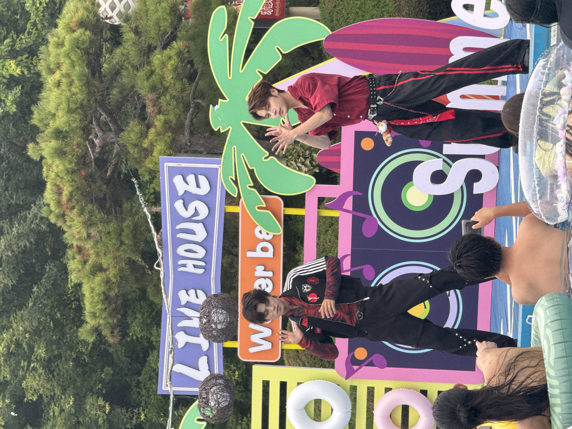 Two young men in trendy outfits are performing on a stage in what appears to be an outdoor event, possibly a water park or festival, given the presence of inflatable rings and people in the water in the foreground. The man on the left is wearing a red and black plaid shirt under a black jacket with white stripes and patches, black pants, and black shoes. He is waving with his right hand, and his left hand is raised. The man on the right is wearing a red short-sleeved button-down shirt, black pants with red stripes and studded details, and black shoes. He has his hands raised in a gesturing motion. Behind them is a colorful backdrop featuring graphics of musical notes, a palm tree, and large white letters spelling "Summer." A purple sign above the stage reads "LIVE HOUSE," and another orange sign says "Water ba." The background consists of lush green trees and foliage. In the foreground, several people are partially visible, seated in inflatable rings in a pool of blue water. One person in the foreground is pointing towards the stage. The scene suggests a lively summer performance event.