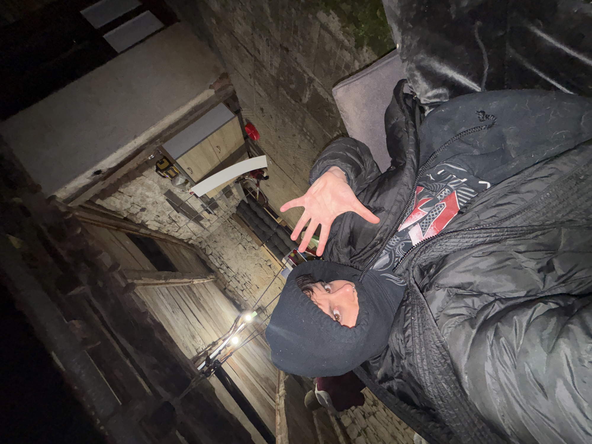 The image, taken at an extreme angle, shows a person in Karlukovo, Bulgaria, lying on a gray couch with their legs near a white cabinet. The person is wearing a black hooded sweatshirt and a black puffer jacket, and their hand is extended towards the camera. The background shows a decaying brick structure with a wooden frame.