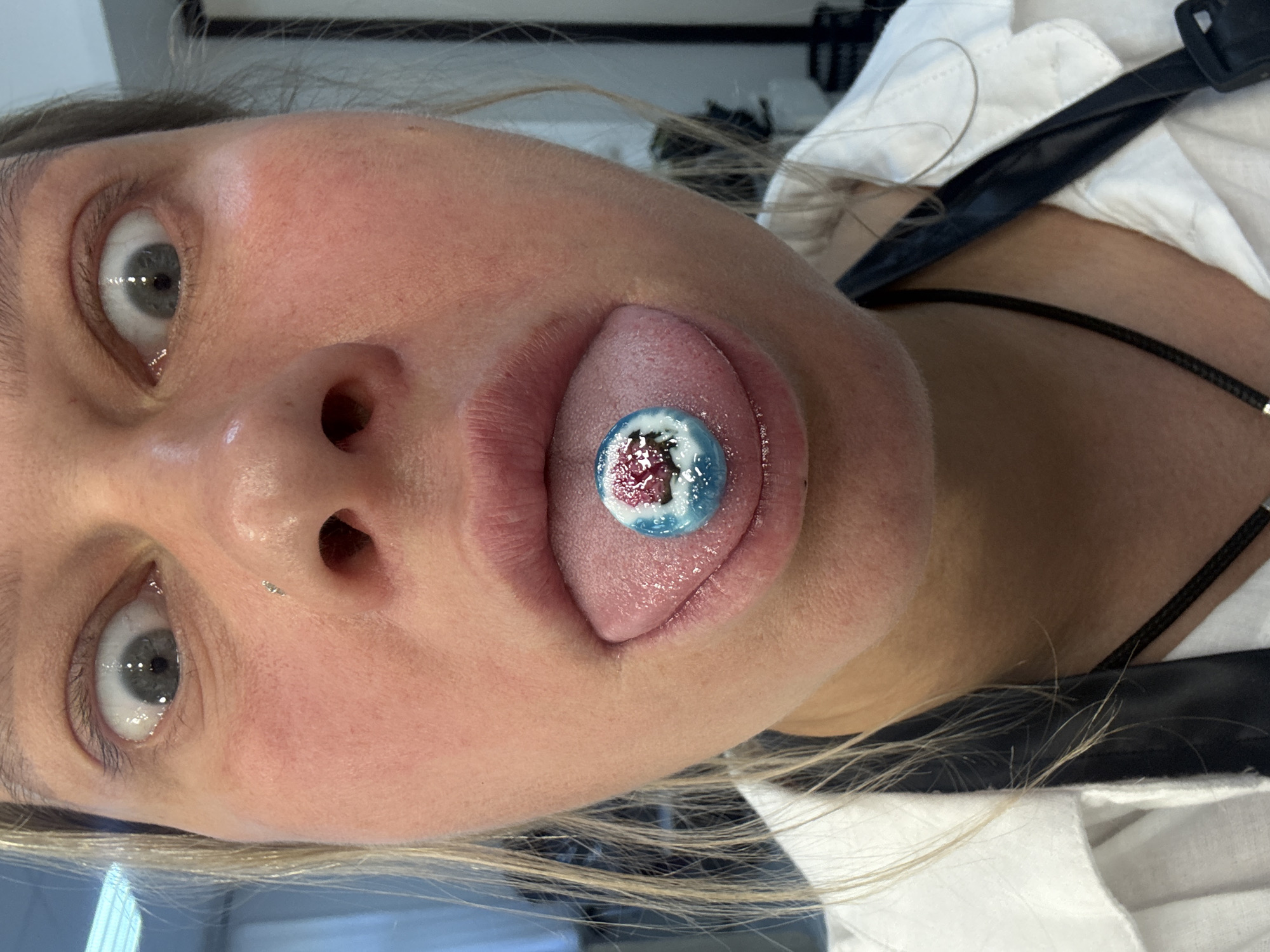 A close-up photograph shows a fair-skinned woman with her tongue extended, holding a small, round object in her mouth. The object appears to be a candy or dessert item, decorated with blue, white, and red colors. The woman has light-colored eyes, and a piercing is visible on her left nostril. She is wearing a white shirt and a black strap. The image is angled, with the woman's face tilted towards the camera. There is no discernible information to identify the location as Perm, Russia.
