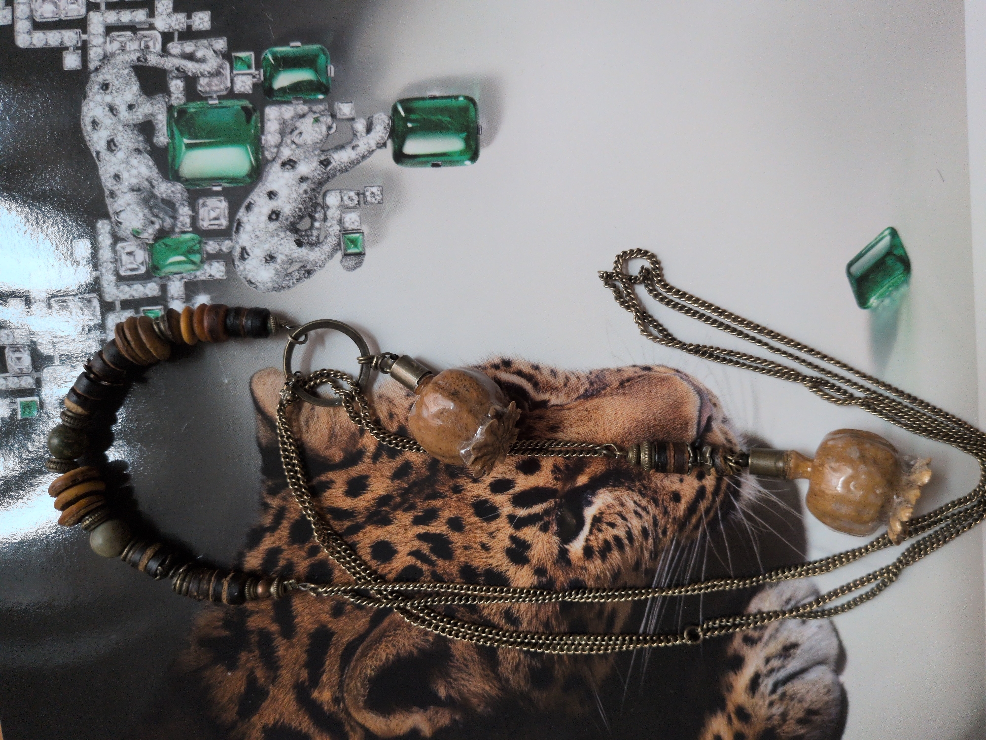 The image displays a close-up of a necklace with multiple strands of aged, brass-colored chain. The necklace features a variety of beads, including dark brown wooden discs, lighter brown wooden discs, and a few greenish-grey spherical beads. Attached to one of the chains is a small, light brown, organic-shaped pendant with a textured surface, resembling a dried fruit or seed pod. Another smaller pendant, cylindrical and made of dark wood and brass, hangs from a shorter chain. The necklace is laid out on what appears to be a glossy magazine or poster featuring a black and brown spotted leopard's face and fur on the left side. On the right side of the poster, there is a stylized illustration of two white leopards surrounded by geometric shapes and emerald green rectangular gemstones. A single, loose, green rectangular gemstone is also present on the white background to the right of the leopard imagery. The scene is lit from the top, casting slight shadows. There is no visible indication of the city of Brandenburg an der Havel, Germany.