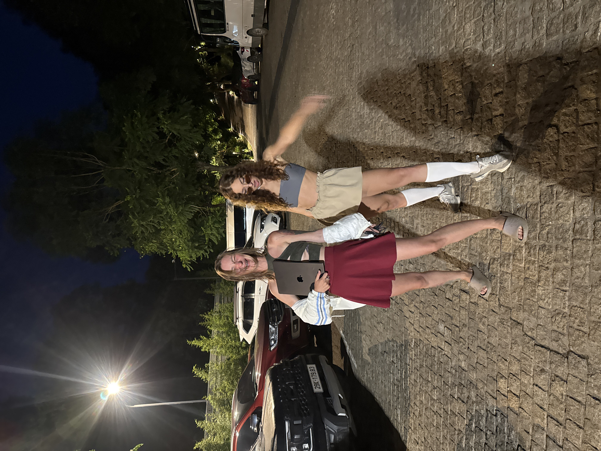 An outdoor, vertical full-shot photograph taken at night captures two young women posing on a paved surface next to parked cars, with trees and a dark sky in the background.
The woman on the left has long, curly brown hair and wears sunglasses, a gray crop top, and light beige shorts. She has white socks and light-colored sneakers, and her right arm is blurred, suggesting a dynamic pose. She has a faint smile.
The woman on the right has long, straight light brown hair and is wearing a dark green tank top and a maroon skirt. She holds a silver laptop (appearing to be a MacBook) in her left arm and wears light grey slides. She is smiling broadly with her mouth open, looking directly at the camera. A white and blue striped jacket is visible slung over her arm or waist.
Behind the women, several vehicles are parked, including a white van/SUV and a prominent dark red pickup truck. A bright streetlight with a starburst effect is visible in the lower left background, casting light on the scene. The ground is paved with rough, irregular stones. Lush green foliage and trees are visible in the background, some illuminated from below.
The scene takes place at night, indicated by the dark sky and artificial lighting. Both women appear happy and relaxed, posing for the camera.
Visible text includes the license plate "29K-179.83" on the red pickup truck, which is a Vietnamese license plate, confirming the location context of Púng Luông, Vietnam.
