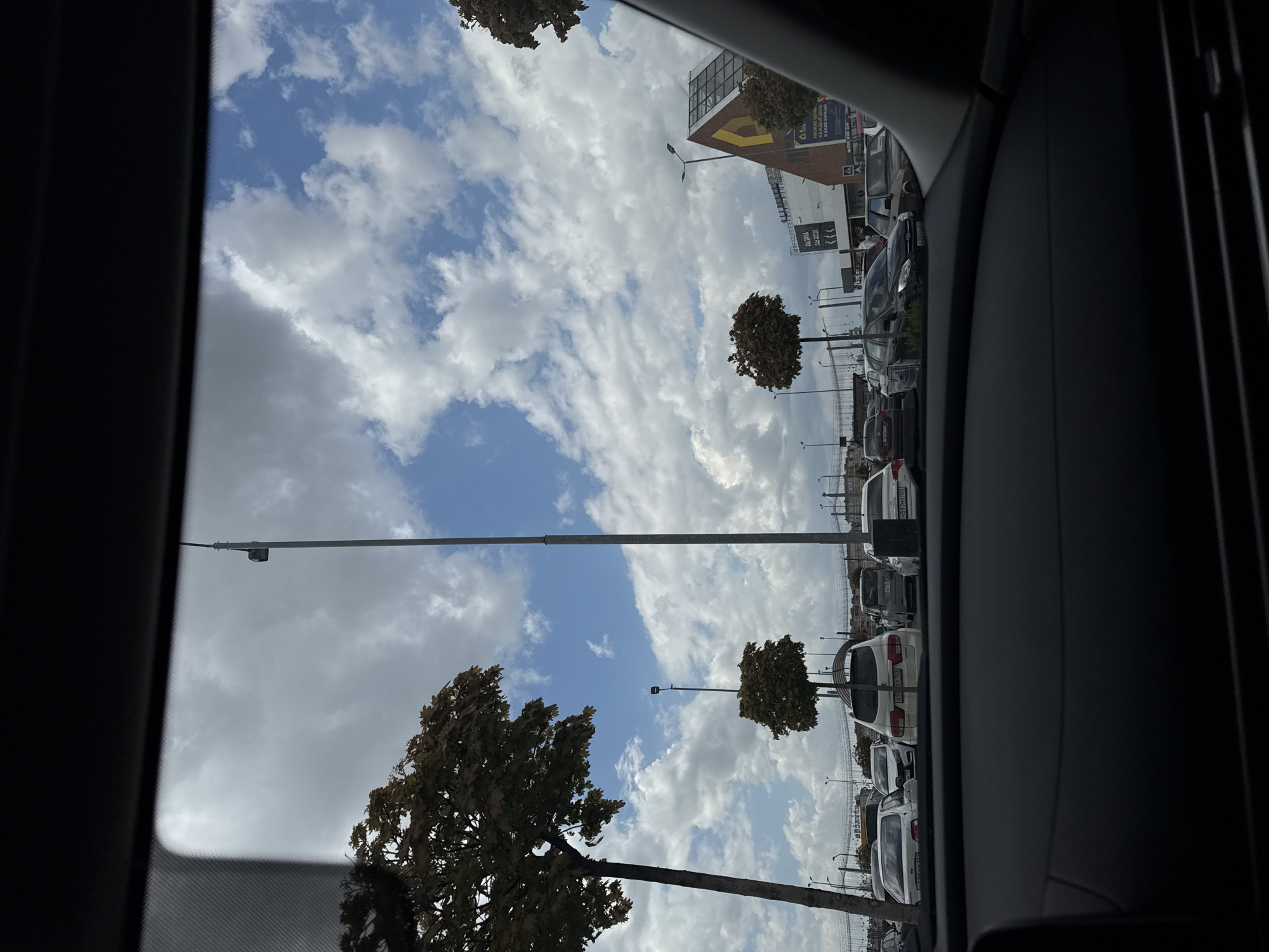 The image shows a view from inside a car, looking out through a window. The outside scene includes a parking lot with rows of parked cars. Several trees are visible in the parking lot, along with street lights. In the background, there is a large building with signs. The sky is partly cloudy, with patches of blue sky visible between the clouds.