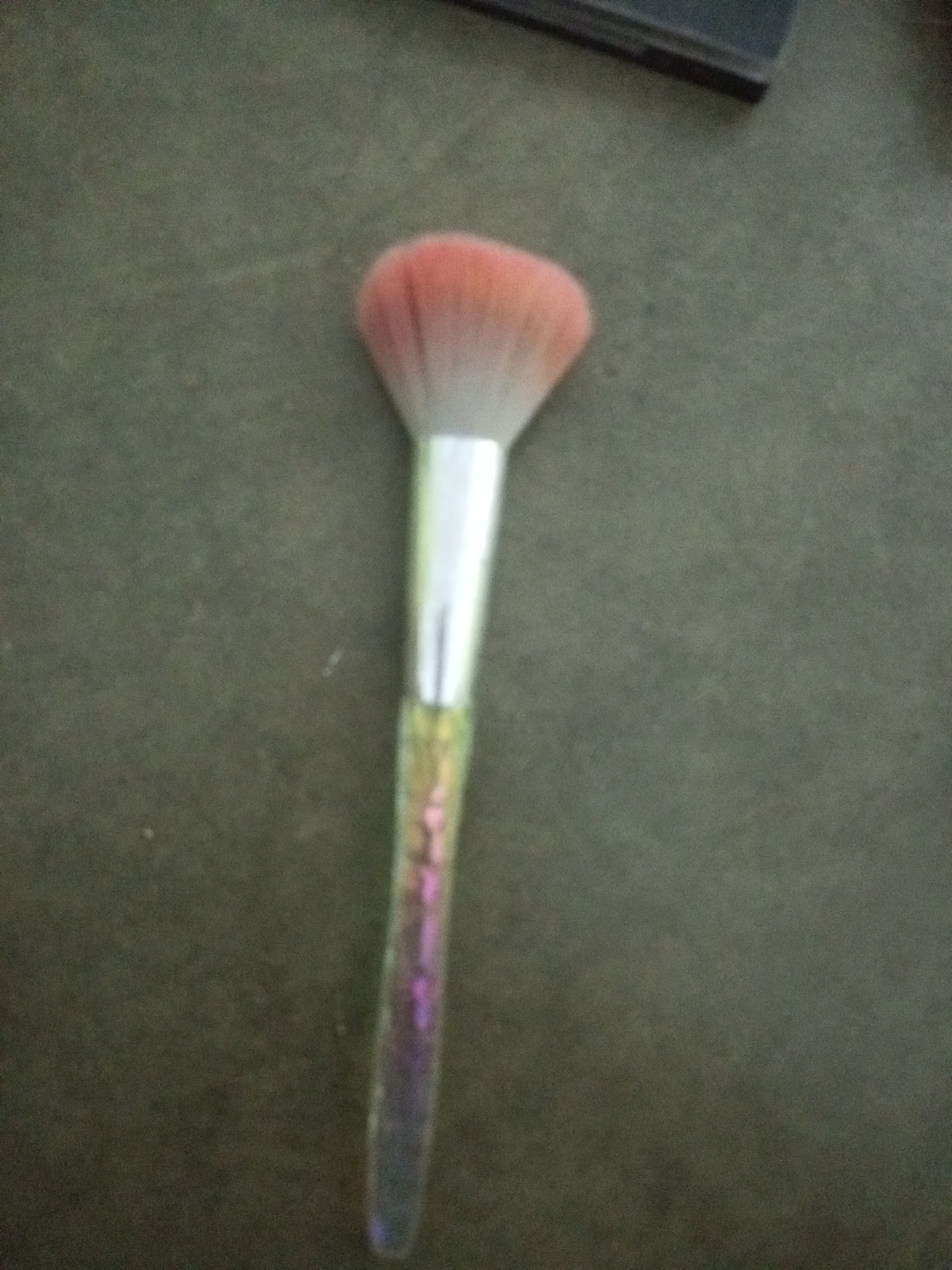 A makeup brush with a fluffy pink tip and a holographic handle rests vertically on a dark, textured surface, possibly a table or floor. The handle exhibits a gradient of colors, shifting through shades of purple, blue, and green. The top of the image shows a blurred dark object, which might be a piece of furniture or electronic equipment. The scene is indoor and dimly lit. There are no people or any other discernible objects in the immediate vicinity. There is no information in the image that can be used to determine its location in La Tagua, Venezuela.