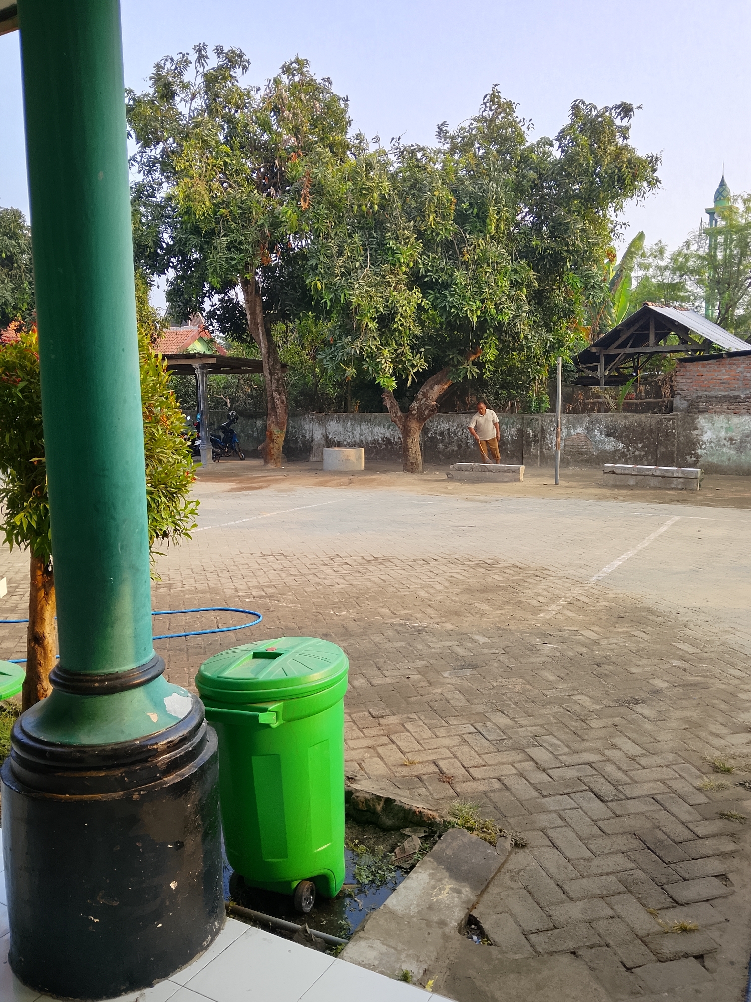 In the city of Kepuh, Indonesia, a daytime outdoor scene unfolds. In the foreground, a large, weathered green pole stands to the left, with a black base. Next to it is a bright green trash can on wheels. The ground is paved with light-colored interlocking bricks. In the middle ground, a paved area, possibly a courtyard or sports court, is visible. A man in a light-colored shirt and brown pants is sweeping the area with a broom. Behind him, a large tree with dense green foliage dominates the background. To the right of the tree, a rudimentary open-sided shelter with a dark roof stands. Further in the background, a brick wall partially obscures buildings and a green minaret of a mosque can be seen against a clear sky. A few motorcycles are parked under a covered area to the left of the sweeping man. The overall environment suggests a community space or schoolyard in a tropical climate.