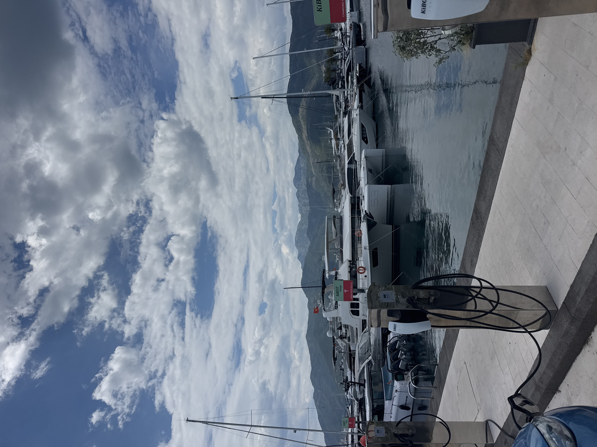 The media file shows a marina with several yachts docked. The scene is set in Tivat, Montenegro, with mountains in the background. The sky is partly cloudy, with patches of blue visible. The water is calm, reflecting the surrounding scenery.
The yachts are of various sizes, with some appearing to be luxury vessels. They are neatly arranged along the dock, with their masts extending upwards. Some of the yachts have their flags flying.
In the foreground, there are charging stations for the boats, with cables coiled neatly. The dock is made of light-colored paving stones. The overall impression is one of a well-maintained and active marina.
The time of day appears to be daytime, given the lighting conditions. There are no people clearly visible in the image.
Visible text includes "KIBO" on a green flag on one of the yachts and "53" on the stern of another yacht. There are also some smaller labels on the charging stations that are difficult to read.