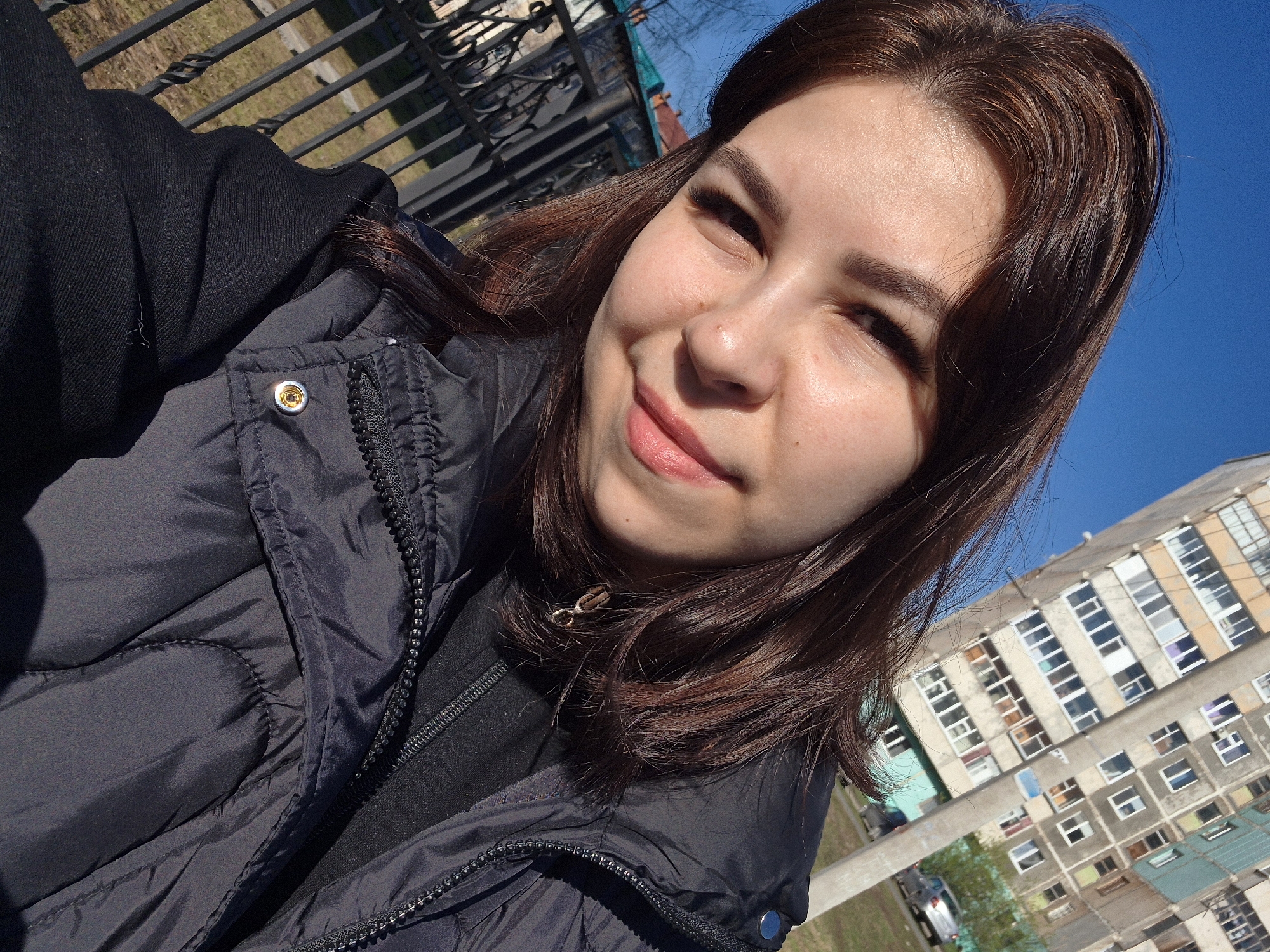A young woman is taking a selfie outdoors on a sunny day. She has brown hair and is wearing a black puffer jacket. The background shows a clear blue sky and a multi-story building. The building appears to be a residential apartment complex with many windows. Given the description of the location being Tobolsk, Russia, it can be assumed that the building is likely a Soviet-era apartment block. The woman is smiling, and the photo is taken from a slightly low angle. A metal fence is visible at the upper-left corner of the image.