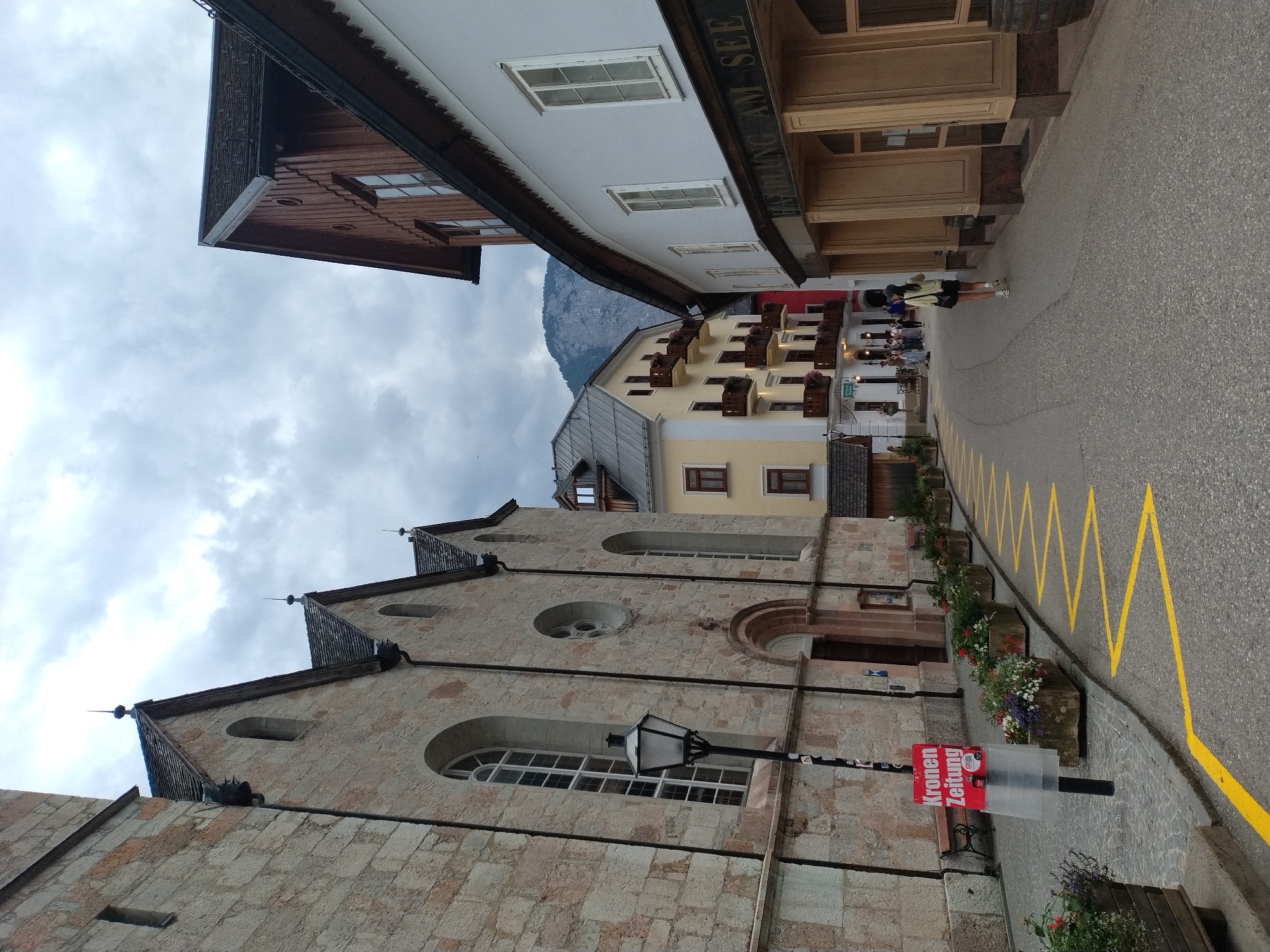 The image shows a street scene in Hallstatt, Austria, dominated by the facade of a stone building on the left, likely a church, with arched windows and a steep roof. A lamppost stands in front of it, and a red sign that reads "Kronen Zeitung" is attached to a pole. A row of stone planters with flowers lines the sidewalk.
The street curves to the right, with buildings of various architectural styles lining its sides. On the right, a white building has arched windows and displays the text "Handlung am See" above its ground-floor arcade. Further down the street, a yellow building with balconies is visible.
The road surface is paved, with yellow zigzag markings painted on it. Several people are walking along the street, and a lone female figure in a light-colored jacket and dark shorts is walking away from the camera towards the right side of the frame. The sky is overcast, and a mountain range is visible in the background. The overall atmosphere is that of a European town on a cloudy day.