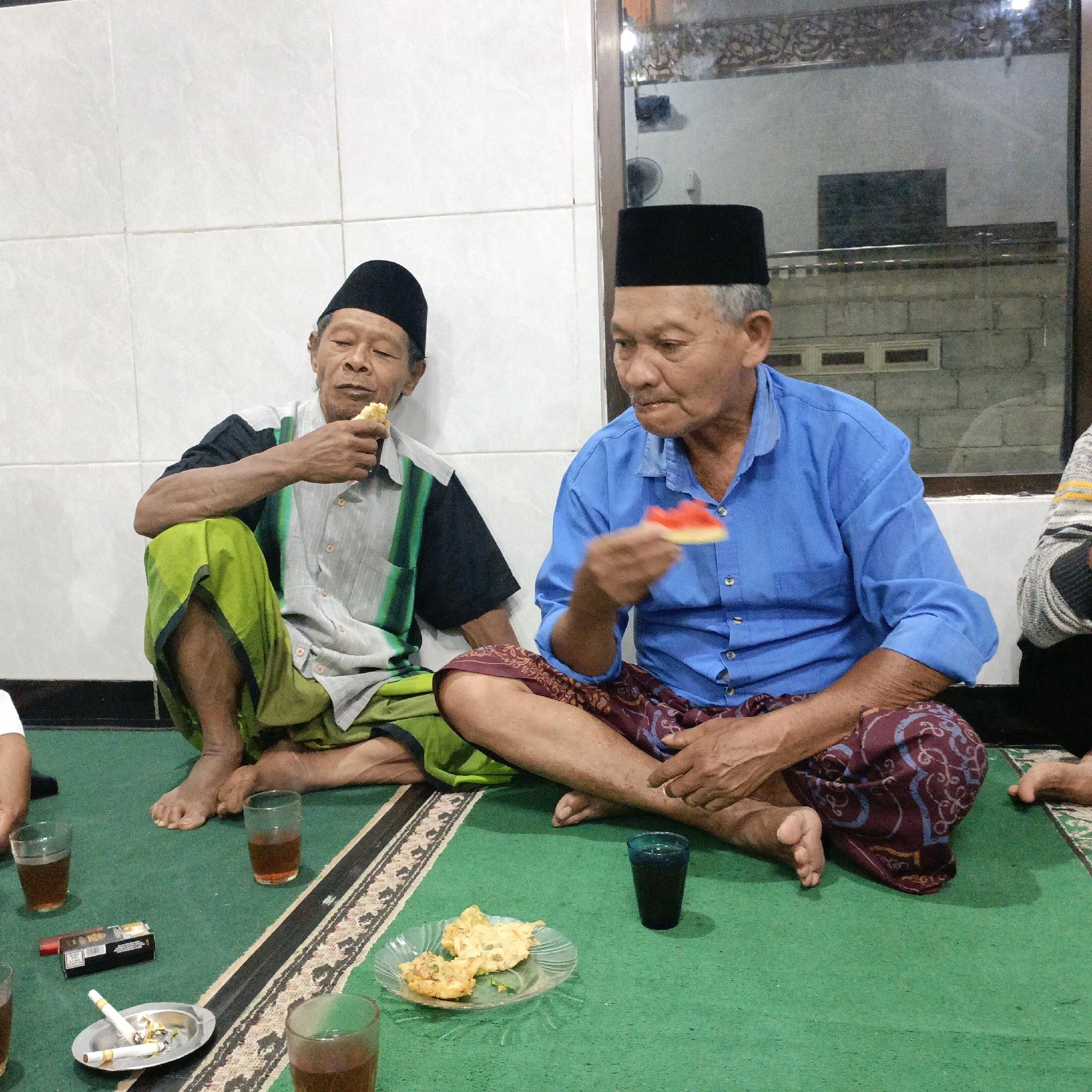 Two elderly Indonesian men, dressed in traditional attire with black peci caps, sit on a green carpet in what appears to be a home in Salatiga, Indonesia. The man on the left, wearing a striped shirt and a green sarong, is eating a piece of food. The man on the right, in a blue shirt and a patterned sarong, holds a small, red object, possibly food, in his hand. Between them, on the carpet, are several glasses of tea, a plate of fried food, a pack of cigarettes, and two lit cigarettes in an ashtray. The background shows a tiled wall and a reflective surface that may be a mirror or window, hinting at an indoor setting. The scene suggests a casual gathering or meal.