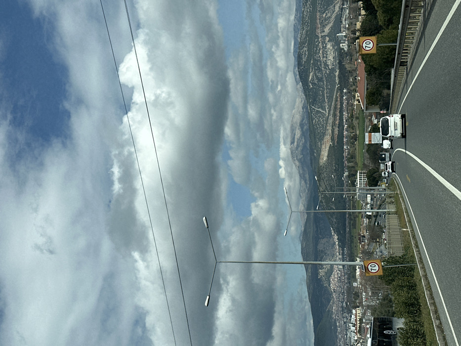 The image displays a daytime view from a vehicle on a multi-lane highway leading towards the city of Menteşe, Turkey. The sky is partially covered by large, grey and white cumulus clouds, with several patches of blue sky visible. Two thin, dark power lines cross the upper portion of the frame.
In the foreground, a paved road with white solid lane markings and a yellow dashed line extends into the distance, curving slightly to the right. A tall, grey streetlight pole with a dual horizontal lamp fixture is prominent on the left side of the road. A yellow circular speed limit sign, featuring a black border and the number '70' in black, is positioned on a pole to the right of the road. Further down the road, a series of similar streetlights recede from view.
Several vehicles are observed on the road, all moving away from the camera. A white panel van occupies the right lane, followed by a white SUV. A dark car and another white vehicle are visible further down. On the far left, a portion of a large, dark vehicle, possibly a bus, is partially obscured behind a concrete barrier that lines the left side of the highway. A metal guardrail runs along the right side.
The midground reveals the urban landscape of Menteşe, characterized by various buildings and interspersed green vegetation. On a hillside to the right, a large quarry or excavation site with exposed rock faces is visible. The background is dominated by a mountain range, with the most distant peaks appearing distinctly snow-capped, while closer mountains are covered with trees and sparse vegetation.