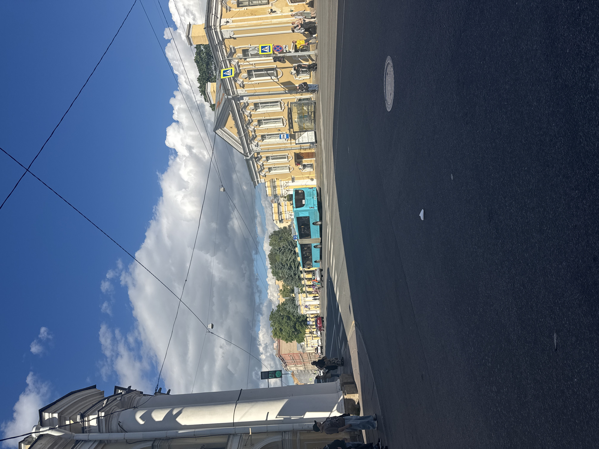The image depicts a street scene in Saint Petersburg, Russia, likely during the daytime with a bright, partly cloudy sky. The perspective is low, looking up at buildings and the sky.
The foreground shows the asphalt street and a sidewalk. Several people are visible on the sidewalk and near the buildings, some standing and others sitting. A blue bus is the most prominent object in the mid-ground, traveling down the street. On the left side of the street, a yellow building with multiple windows and architectural details dominates the scene. Pedestrian crossing signs and traffic lights are also visible near the building. Further down the street, other buildings and trees line the road.
The scene appears to be a typical urban environment. The bright sky suggests fair weather, though scattered clouds indicate the possibility of changing conditions. The presence of a bus, pedestrians, and street infrastructure points to a public street in an active part of the city. No specific event is evident, but the overall impression is one of daily life in a European city. No discernable text is visible in the image.