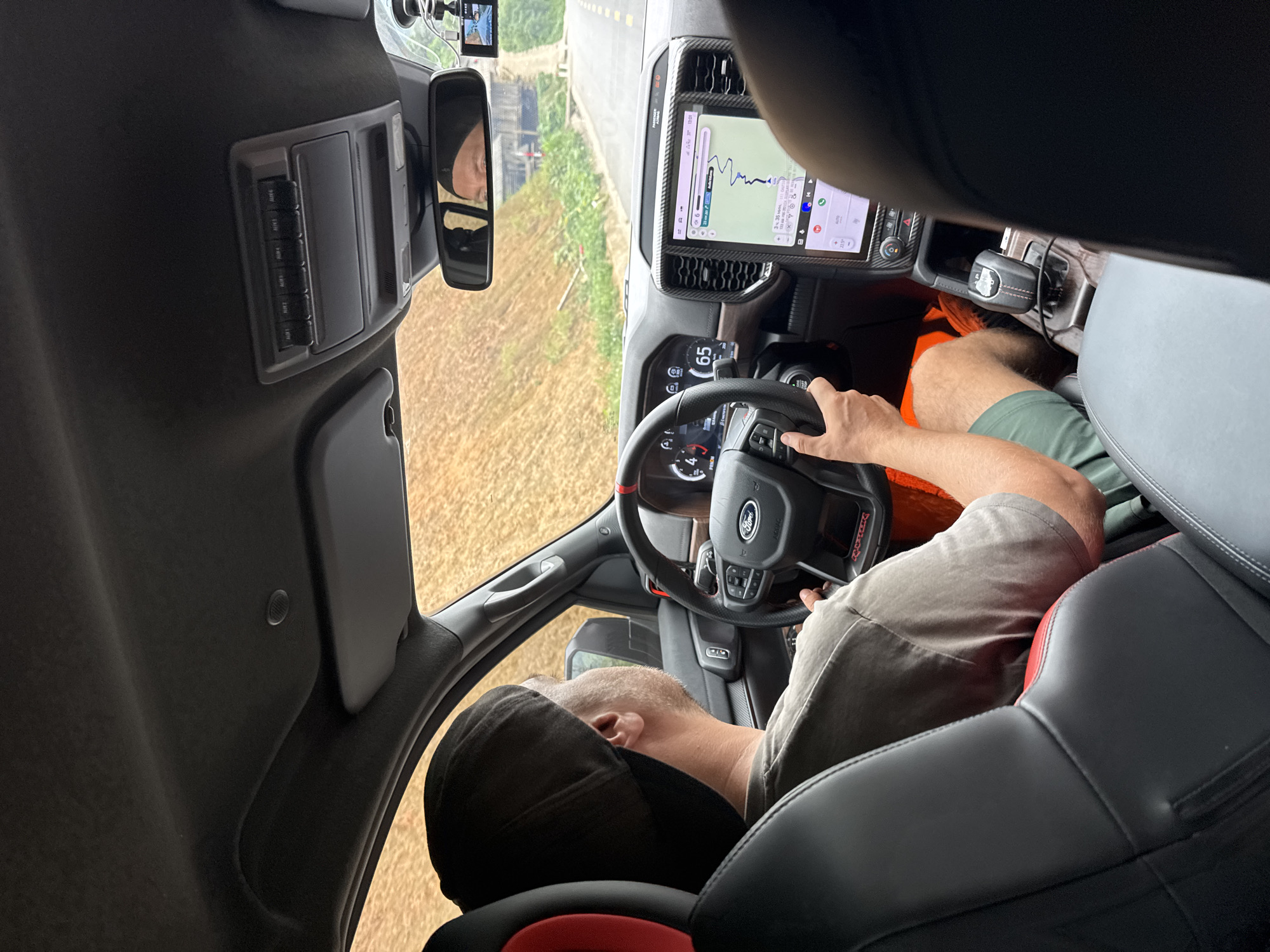 The media file captures the interior of a vehicle from a high angle, looking forward from the rear passenger side. A male driver, visible from behind, wears a black cap, a grey t-shirt, and green shorts. His left hand rests on the steering wheel, and his reflection is faintly visible in the rearview mirror.
The vehicle's interior features a large, vertical infotainment screen displaying a navigation map with a route line and an elevation profile, showing the time "13:01" and a temperature of "22.5°". Below the screen, the dashboard and center console are visible, including a rotary gear shifter with "TRX" stitched in red on its boot. A small screen behind the steering wheel shows the speed as "65". An overhead console above the rearview mirror contains six auxiliary switches labeled "AUX 1" through "AUX 6". A small camera or device is mounted on the windshield. The steering wheel, however, clearly features a "Ford" logo and "Raptor" text, creating conflicting brand identifiers within the vehicle's cabin which otherwise strongly suggests a Ram TRX due to the large screen and shifter design.
Outside the car, a road is visible ahead, bordered by green and brown vegetation, consistent with a rural or semi-rural landscape. The scene indicates bright daylight and clear weather. The overall activity is driving and navigation, likely within Minh Sơn Commune, Vietnam, given the contextual information. No specific emotions are discernible from the partially obscured driver.