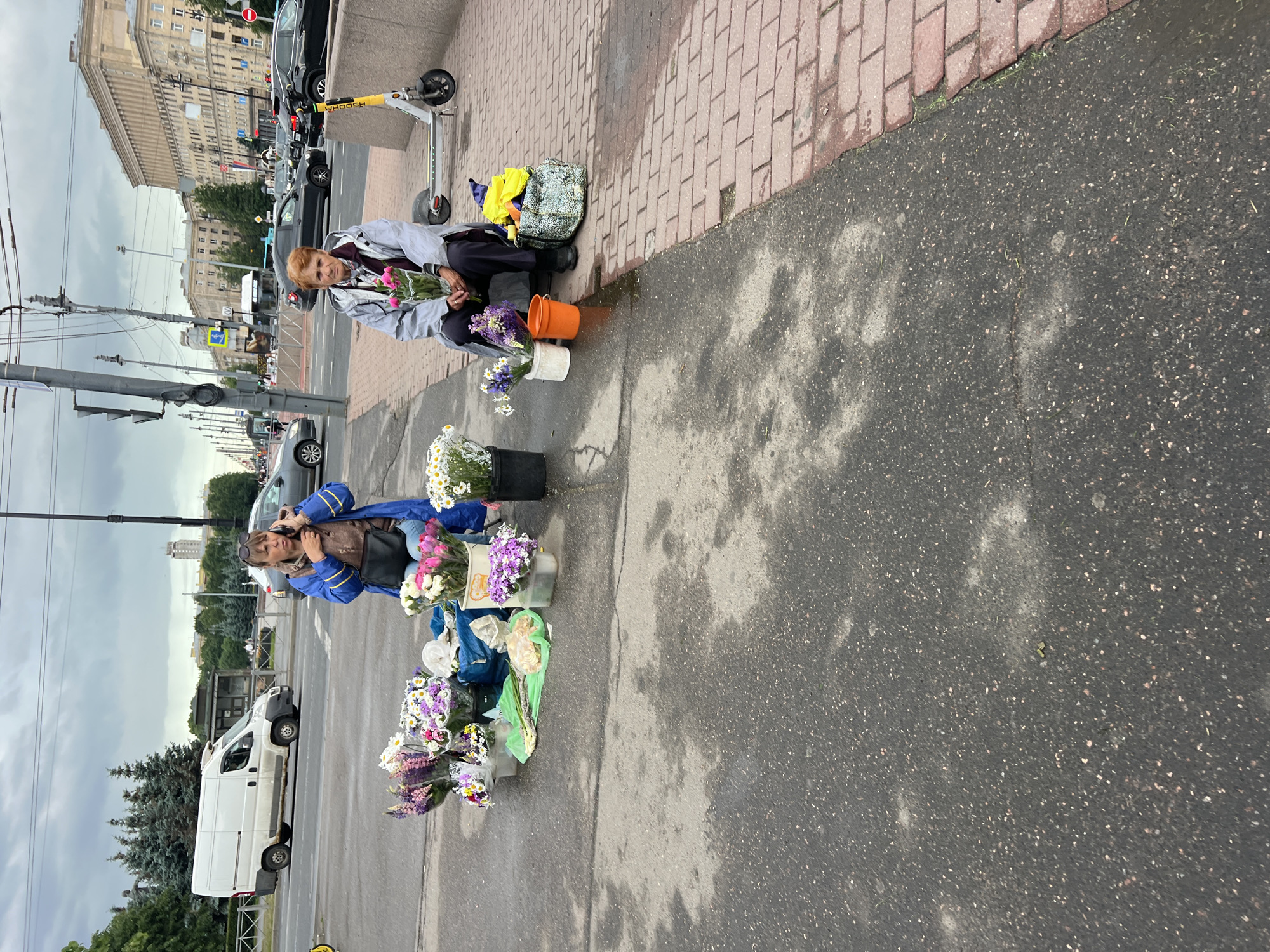 This image depicts a street scene in Saint Petersburg, Russia, under an overcast sky. Two women are present, engaged in selling flowers. The woman on the left, wearing a blue jacket and jeans, is standing and speaking on a mobile phone. She is positioned behind a collection of assorted flowers in buckets and bags. The woman on the right, wearing a grey jacket and dark pants, is sitting on a curb. She is holding a bouquet of pink flowers and has a shopping bag and an orange bucket beside her. A yellow electric scooter is parked behind her.
The setting is a roadside area with a paved sidewalk made of red bricks and an asphalt road. In the background, traffic is visible, including cars and a white van. Buildings with distinctive architectural features, typical of Saint Petersburg, line the street. Trees and streetlights are also present. The overall atmosphere appears to be a typical urban street environment during the day.