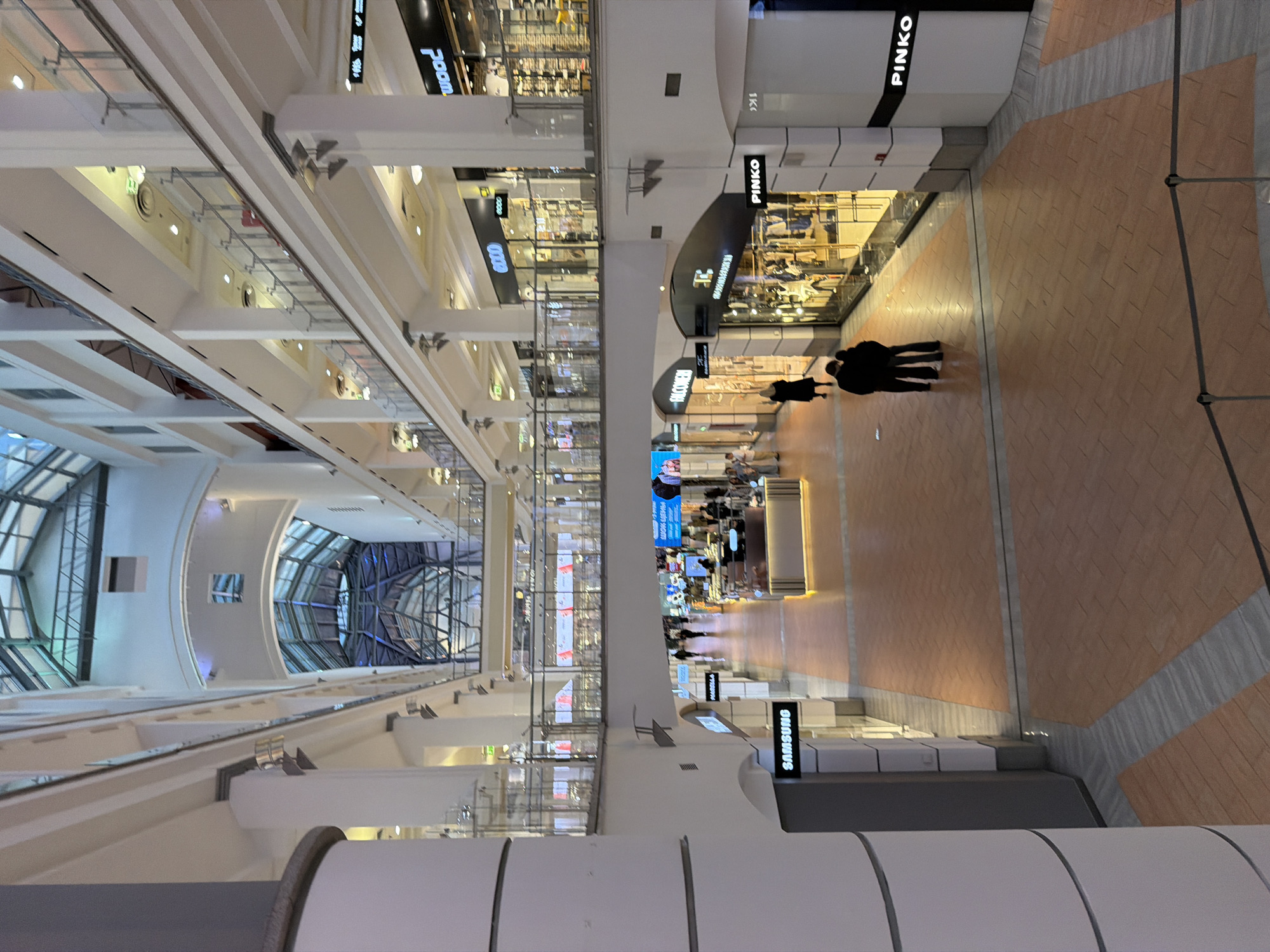 This image depicts the interior of a shopping mall, likely in Unknown, Ukraine. The architectural style is modern, with high ceilings and glass panels. A distinctive feature is the arched glass roof visible in the distance. The mall has multiple levels, with shops lining the walkways. Several signs for known brands, like PINKO and SAMSUNG, are visible. Several people are present. The lighting is bright and appears to be artificial. The floor is covered with a patterned tile. The angle of the shot is upward, seemingly from a lower level looking up and along the length of the mall.