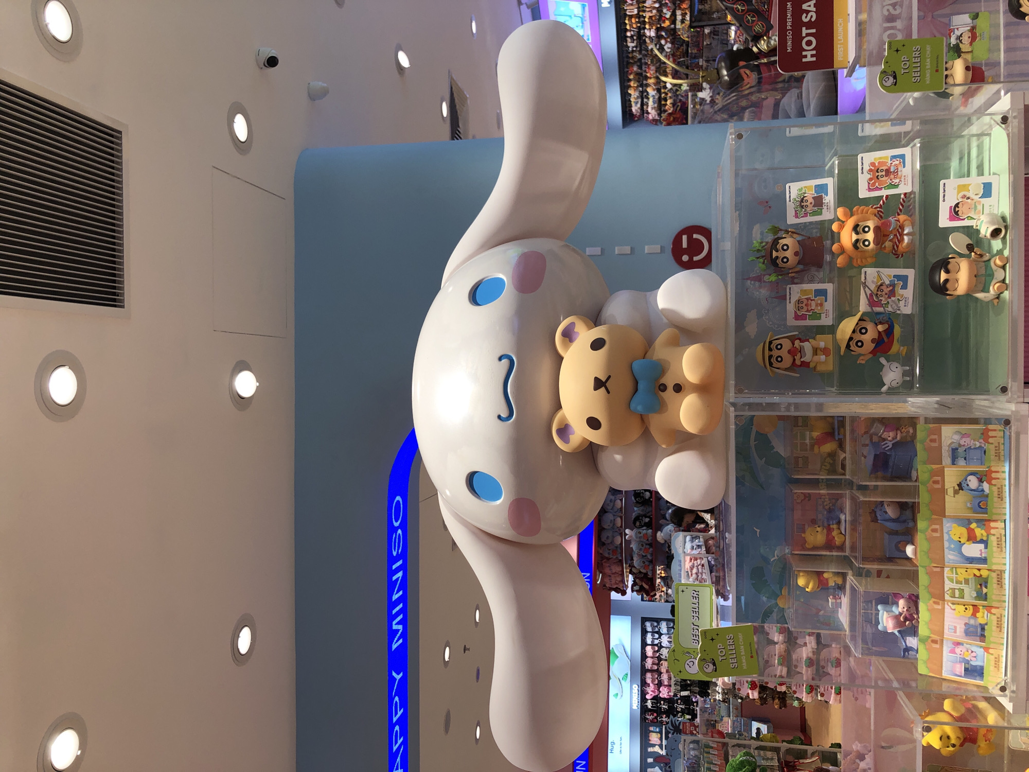 This media file captures a brightly lit indoor scene, likely a retail store, with a prominent, oversized cartoon character sculpture of a white rabbit with large blue eyes and pink cheeks, holding a teddy bear. The character is situated in the foreground and appears to be the central attraction. Behind the sculpture, shelves filled with various merchandise, including small figures and plush toys, are visible, suggesting a toy or character goods store.
Several prominent signs display text, including "MINISO PREMIUM HOT SALE FIRST LAUNCH," "BEST SELLER," and "TOP SELLERS HÀNG BÁN CHẠY." The "MINISO" branding suggests the store is part of this popular retail chain. The overall atmosphere is cheerful and engaging, typical of a family-friendly shopping environment. The lighting is bright and even, indicating it's likely daytime, though the indoor setting makes it independent of natural light conditions. The presence of security cameras on the ceiling hints at a modern retail setup. The orientation of the image is portrait, with the main character's head towards the top. There are no people clearly visible in the frame, allowing the focus to remain on the display and merchandise.