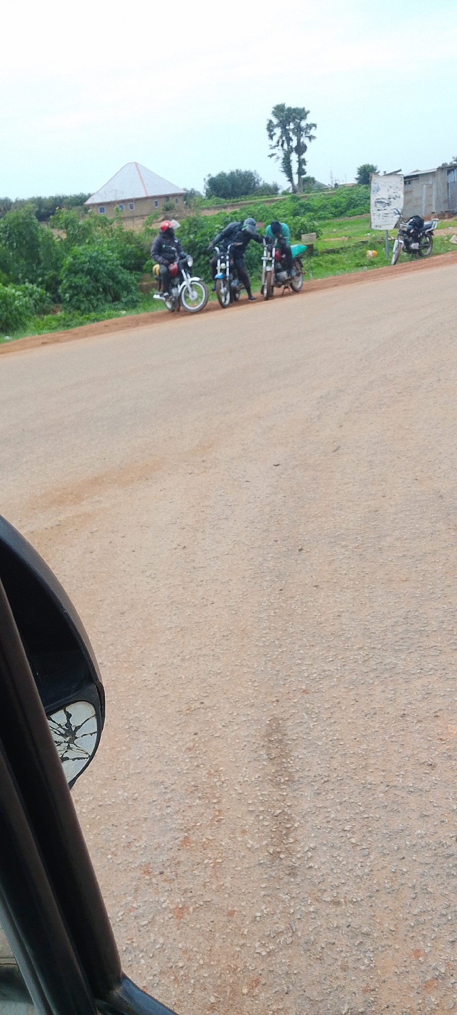 An outdoor, ground-level shot depicts a group of motorcyclists and their motorcycles gathered on a dusty, dirt road. In the foreground, a dark, possibly black, vehicle side mirror is visible on the left side of the frame, its glass cracked. The road extends into the mid-ground where four motorcycles are parked, some with riders. The first motorcycle on the left has a rider wearing a red helmet and a dark jacket. Next to it, another motorcycle is being attended to by two individuals, one wearing a dark jacket and a grey cap, and the other a dark jacket and a green shirt. Further to the right, another motorcycle is parked, and behind it, a fourth motorcycle is visible. The background features a landscape with green foliage and trees, a building with a red-roofed structure, and a tall, slender tree. The sky is overcast.