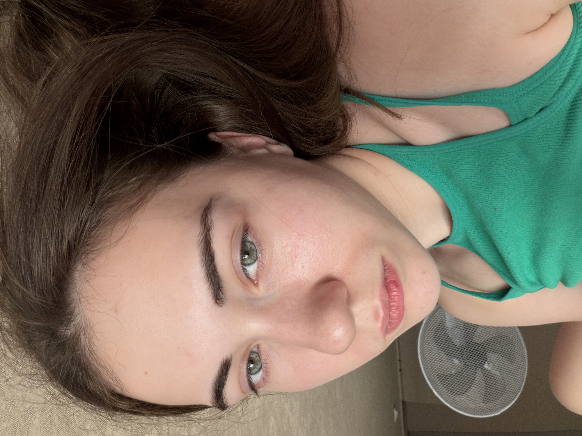 The image shows a close-up, eye-level, horizontal shot of a young woman lying down. She is wearing a green tank top. Her head is tilted to the right, with her face filling most of the frame. She has fair skin, green eyes, and dark brown hair. Her lips are slightly parted. In the background, out of focus, is a white electric fan. The setting appears to be indoors, possibly a bedroom or living area, with a textured, light-colored wall visible. There are no discernible landmarks or elements to suggest a specific location within the city of Tyumen, Russia. The overall impression is that of a casual, personal moment captured in a private space.