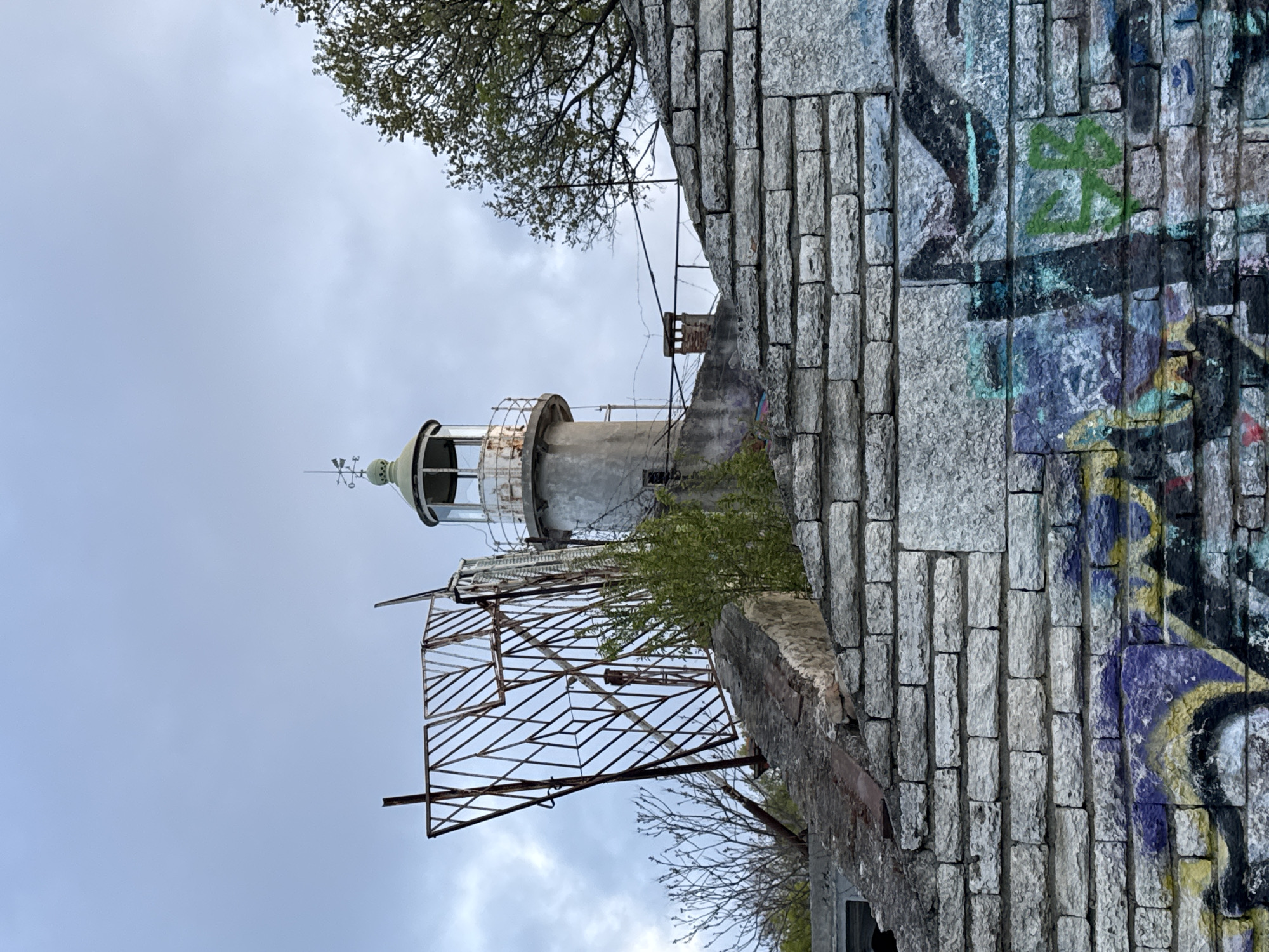 The image depicts an old lighthouse in Varna, Bulgaria. The structure is cylindrical and painted white, with a visible lantern room at the top containing a navigational light. The lighthouse shows signs of decay, with rust and peeling paint. It's partly obscured by vegetation. In the foreground, there's a metal grid structure attached to a brick wall covered with graffiti. The sky is overcast.
