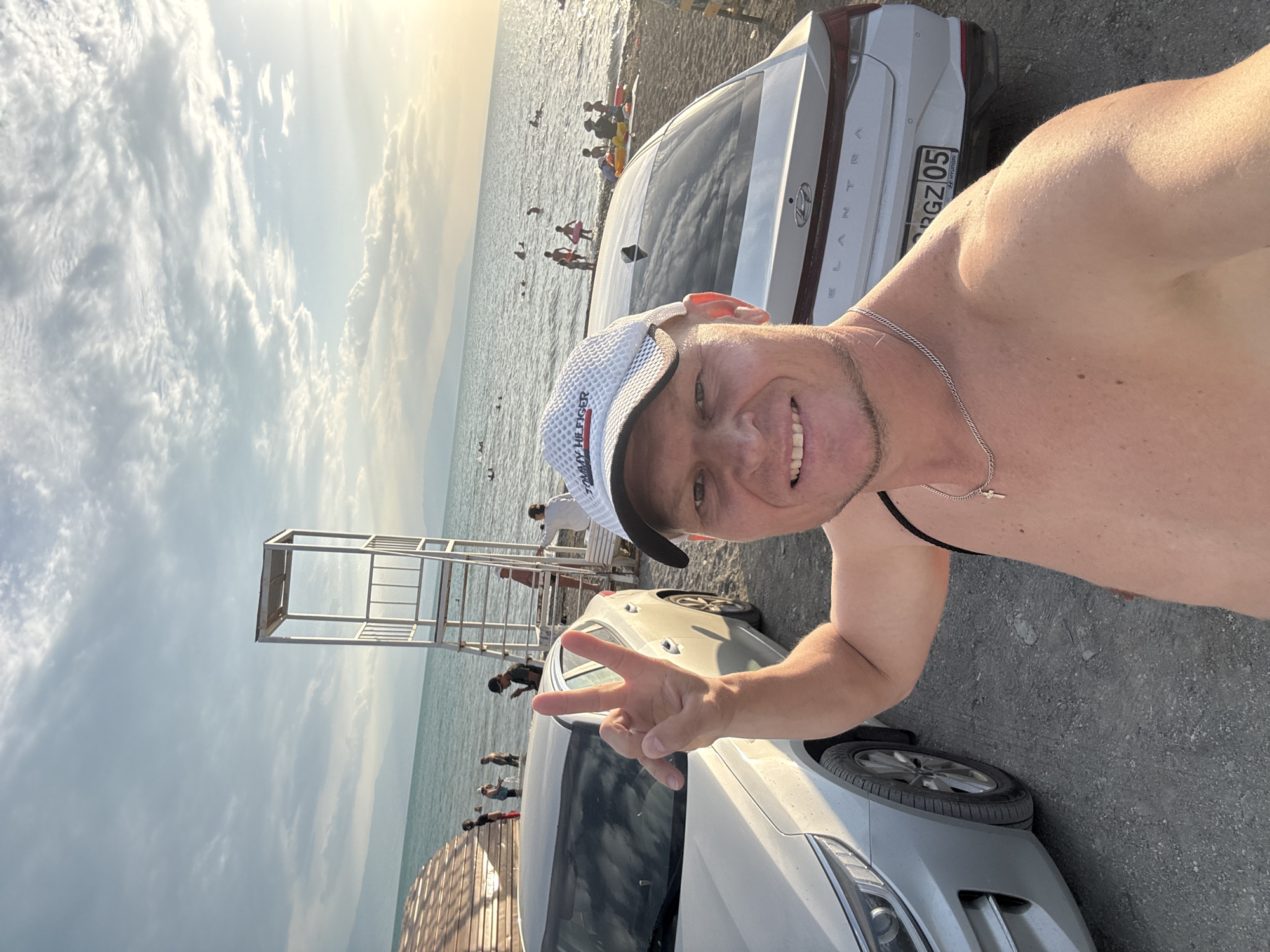 A shirtless man wearing a white Tommy Hilfiger baseball cap and a silver chain with a cross pendant is taking a selfie at a beach. He is smiling and holding up two fingers in a peace sign with his left hand. Behind him, two white cars are parked on a gravelly shore. The man is standing in front of a light-colored sedan, and a white Hyundai Elantra is to his right. The sea is visible in the background with several people swimming and standing in the shallow water. A lifeguard tower stands on the beach. The sky is partly cloudy with the sun setting on the horizon, casting a warm glow. The location is the shore of a body of water, possibly a lake or sea, near the city of Zharbulak, Kazakhstan.