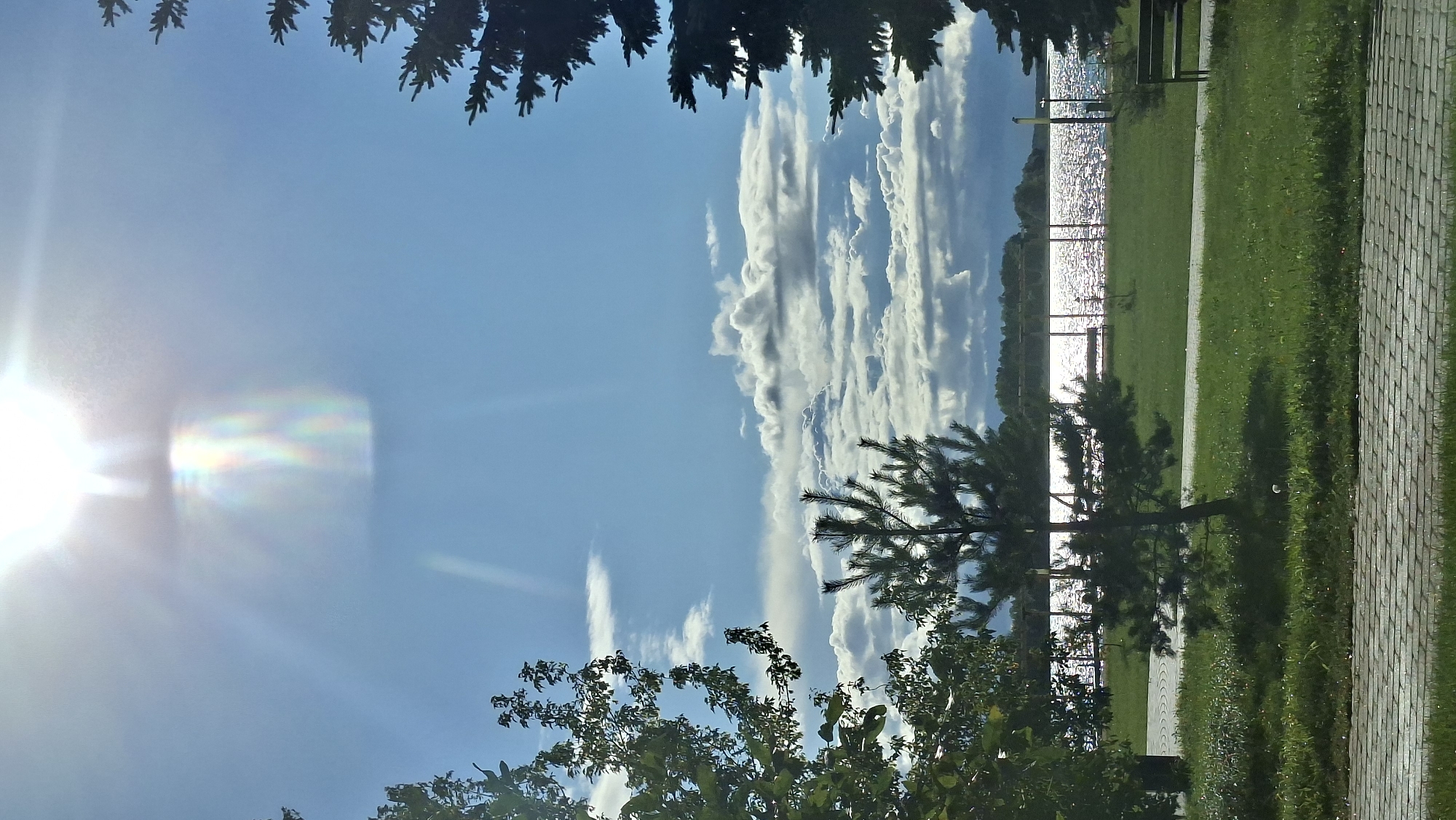 This vertical, full-shot, eye-level, outdoor photograph captures a scenic view of the Myadzyel Reservoir in Myadzyel, Belarus, on a bright, sunny day. The sun is prominently visible in the upper center of the frame, casting lens flares and creating a rainbow-like effect in the sky. The sky is a vibrant blue, interspersed with scattered white and gray clouds, some of which appear dense and dramatic.
In the foreground, the image features a paved walkway or promenade constructed of light-colored rectangular stones. To the left and right edges of the frame, evergreen trees are visible, their dark green needles silhouetted against the bright sky. In the mid-ground, a lush green lawn extends towards the reservoir. A small evergreen tree stands in the middle of the lawn, casting a shadow. Park benches are sparsely placed on the grass, one to the left of the small tree and another to the right, near the edge of the frame.
The Myadzyel Reservoir dominates the background. Its surface sparkles intensely with sunlight, reflecting the sky and clouds. A fence or railing runs along the edge of the reservoir, and beyond it, a treeline of deciduous trees marks the opposite shore, stretching across the horizon. The overall scene conveys a sense of tranquility and natural beauty, characteristic of a park or recreational area near a body of water. There are no people visible in the photograph.