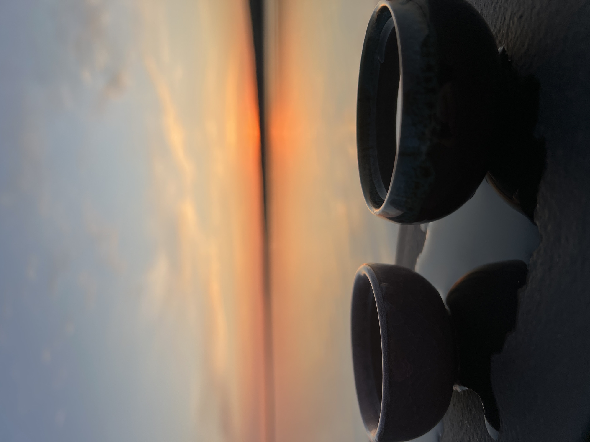 The image, oriented horizontally, depicts two dark-colored ceramic bowls resting on a flat surface. The bowls are stacked vertically, with the top bowl slightly angled compared to the bottom one. The left side of the image showcases a blurred sunset over what appears to be water. The light from the sunset is reflected in the ceramic material of the bowls. Given the location of Chaikovsky, Russia, the body of water is most likely the Kama River. The time of day is likely sunrise or sunset. The overall composition suggests a contemplative or artistic focus.