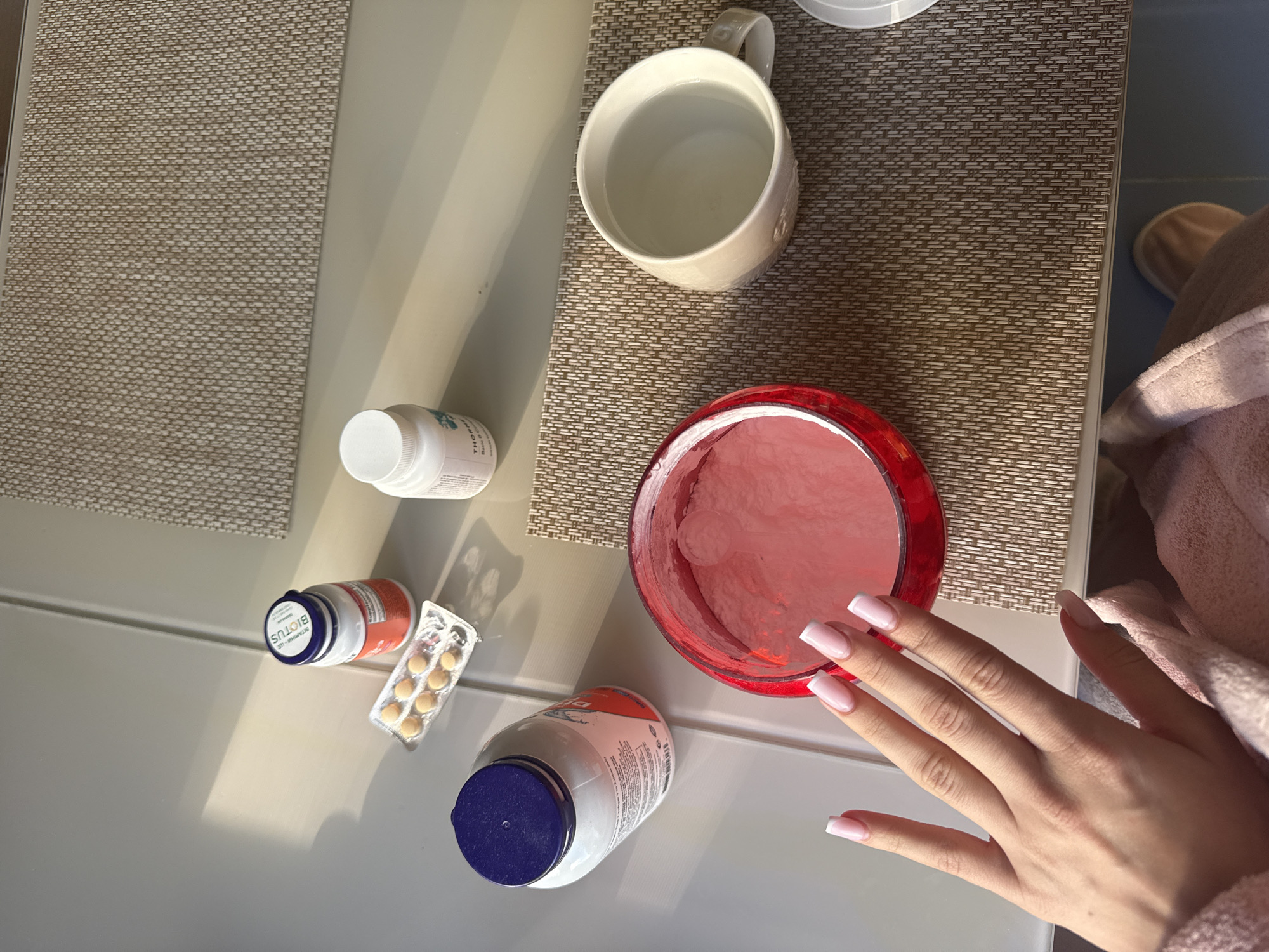 The scene depicts a tabletop setting with several items arranged on it. There are three pill bottles with white and blue caps, a blister pack of pills, a white bottle with a white cap, a red bowl containing a pink substance, and a white mug filled with water. A hand with long, manicured nails is also visible. The tabletop is covered with two different textures: a woven, beige mat under the mug and bowl, and a smooth, light-colored surface elsewhere. A pink bathrobe is partially visible in the bottom right corner. There are no definitive clues in the image to confirm the location as Kyiv, Ukraine.