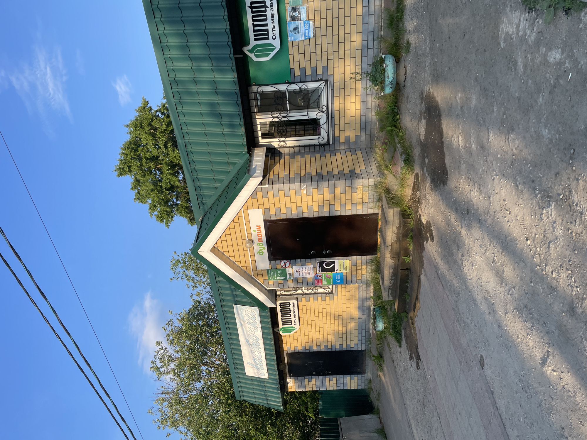 The image shows a small brick building with a green metal roof, likely a storefront, on a sunny day in the city of Prudý, Russia. The building has two entrances, one on the left and one on the right, both with dark brown doors. Above the left entrance, a sign reads "Ласточка" in a cursive font. To the right of this entrance, a smaller sign with a green logo and the text "Штофф" and "Сеть магазинов" is visible. The right entrance is flanked by a window with decorative metal bars and a sign above it that reads "ФуДмаим". Various posters and notices are affixed to the wall to the right of the window. The building is surrounded by sparse vegetation, including a potted plant in front of the right window and some grass and weeds along the facade. The ground in front of the building is a mix of pavement and dirt, with tire tracks visible in the dirt. Overhead, power lines stretch across a clear blue sky. The sun casts long shadows across the ground, indicating it is likely late afternoon or early morning.