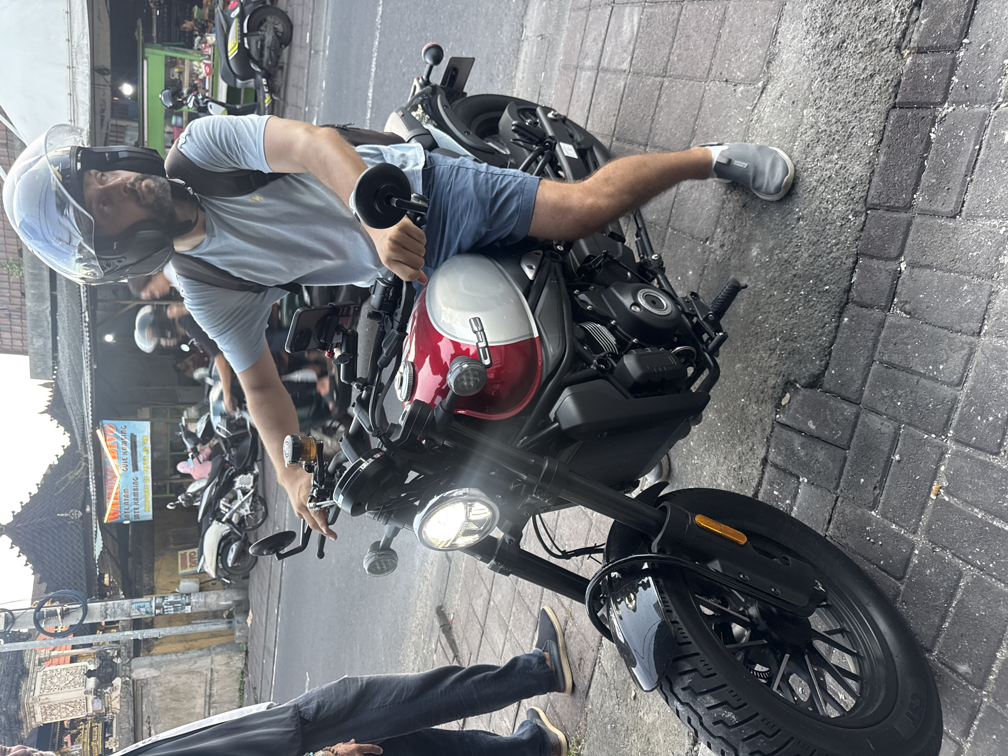 A man with a beard and wearing a full-face helmet, a light blue t-shirt, and blue shorts is straddling a red and white motorcycle in Kuta Selatan, Indonesia. He has one foot on the ground and the other on the motorcycle seat. Another person is standing near the front tire of the bike, wearing blue jeans and brown shoes. The motorcycle is on a brick-paved road. Several other motorcycles are parked in the background. Across the street is a building with a sign that reads "WARUNG GUE KAMBING" along with some other Indonesian text. The sky appears overcast.