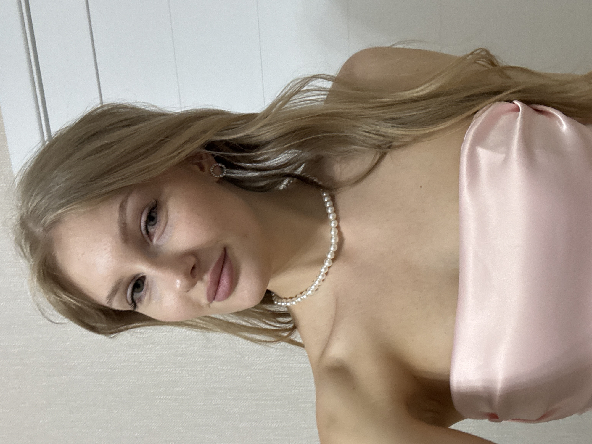 A woman is photographed indoors against a light-colored wall. She is wearing a light pink, strapless garment and a pearl necklace. She has blonde hair, light skin, and light-colored eyes. Her left ear is visible, adorned with an earring. No location is indicated as being in the city of Perm, Russia. The woman is facing the camera and slightly turned to her left.