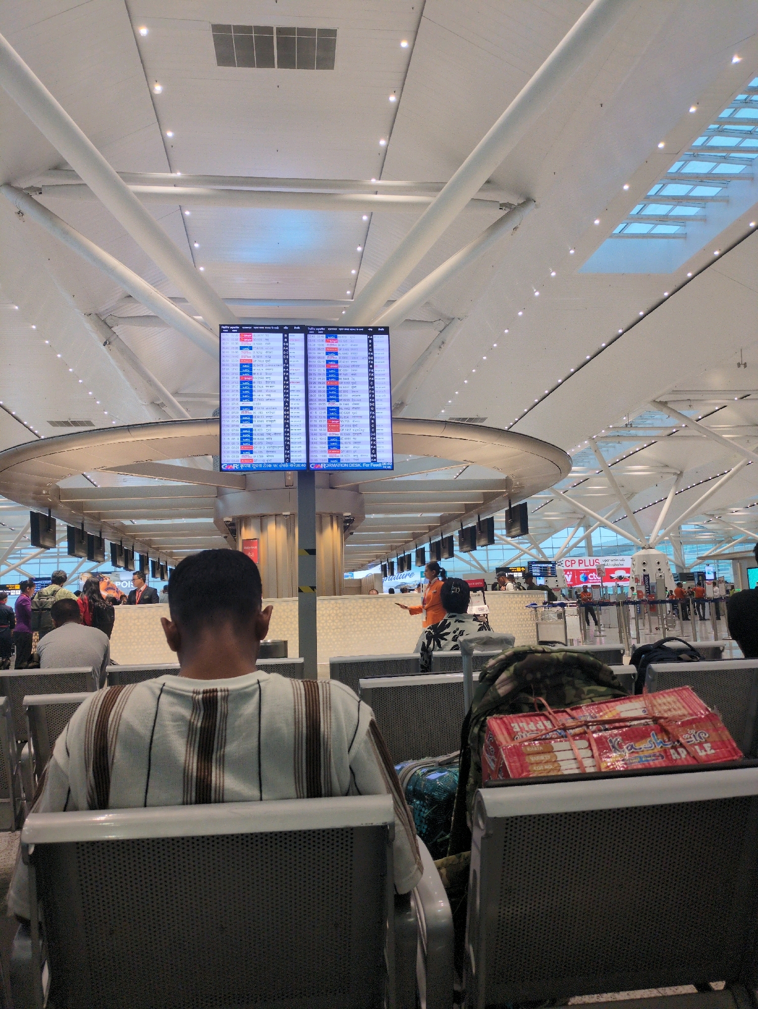 An overhead shot captures a busy airport waiting area in Delhi Cantonment, India. A digital departure board displays flight information in Hindi and English. In the foreground, a person sits with their back to the camera, wearing a striped shirt. Luggage, including a backpack and several boxes of snacks, is placed on the seat next to them. Other travelers are visible in the background, some standing and others seated, interacting with staff or waiting for their flights. The airport's architecture features high ceilings with exposed beams and lighting.