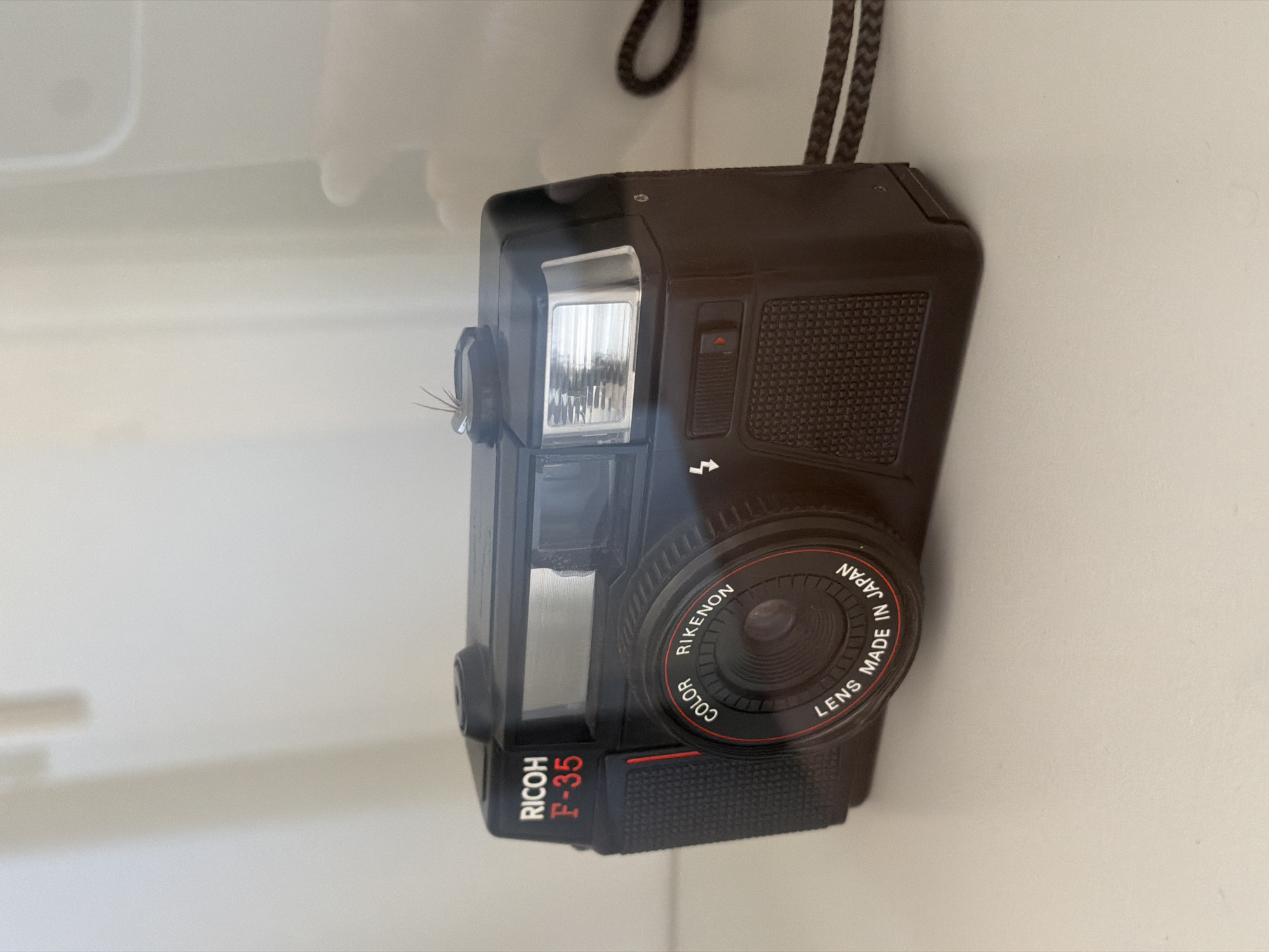 A black RICOH F-35 camera with a silver flash is on a white surface. Text on the camera reads "RICOH F-35" in red and white, and "COLOR RIKENON LENS MADE IN JAPAN" around the lens. There is a brown wrist strap attached to the side of the camera. The background is a white wall with soft shadows. The scene is not in Barcelona, Spain.