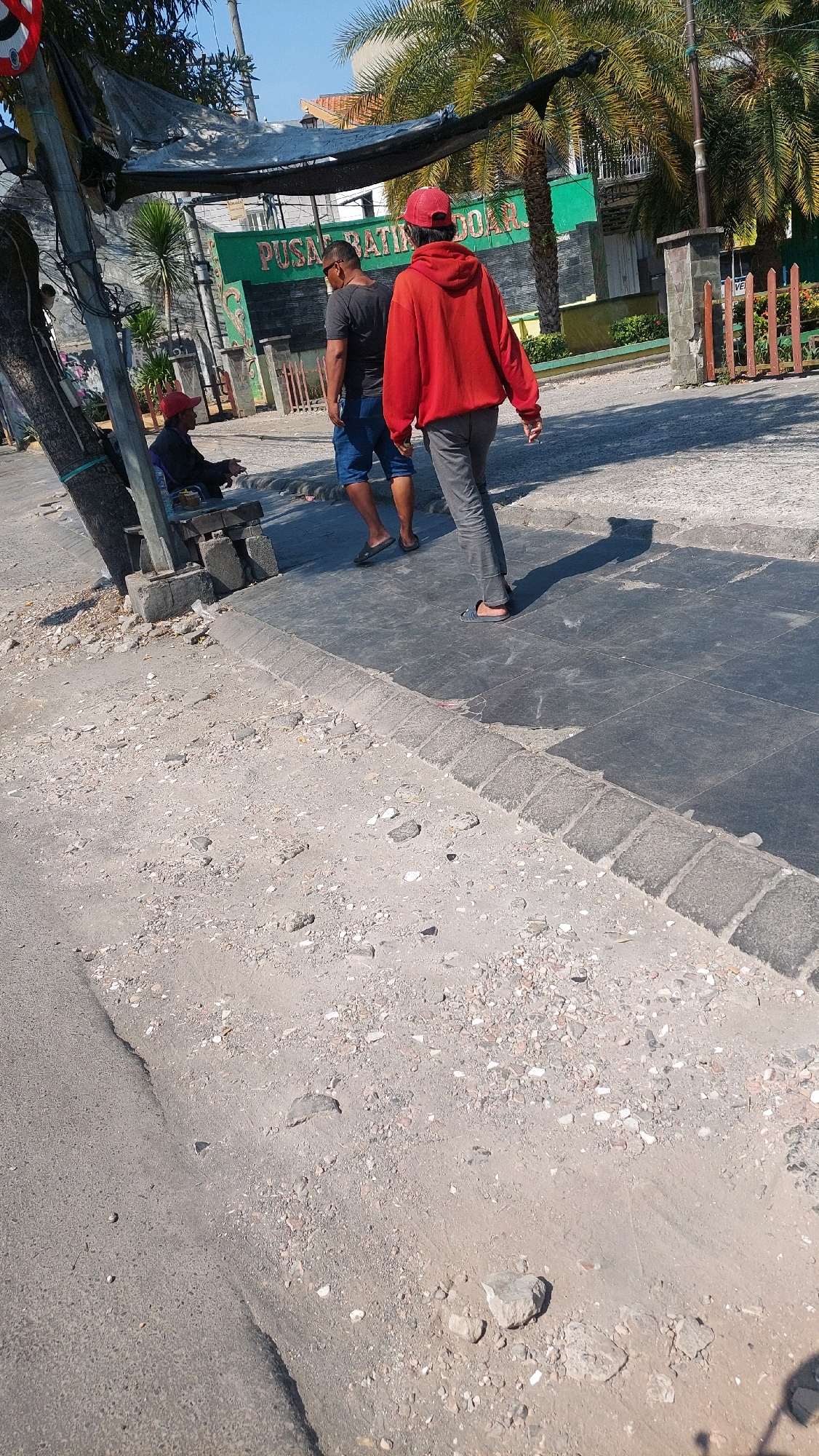 The image depicts a street scene in Sidoarjo, Indonesia. In the foreground, the ground is uneven and covered with dirt and debris. To the left, a person wearing a red cap is seated on a bench under a makeshift shelter. Further back, two men are walking. One man, wearing a red hoodie and a red cap, is walking away from the camera. The other man, wearing a dark t-shirt and blue shorts, is walking alongside him. Behind them, a green building with the text "PUSAT BATIK DOAR" is visible, suggesting it might be a batik center. Palm trees are present in the background, and a wooden fence can be seen on the right side of the image. The sky is clear and blue.