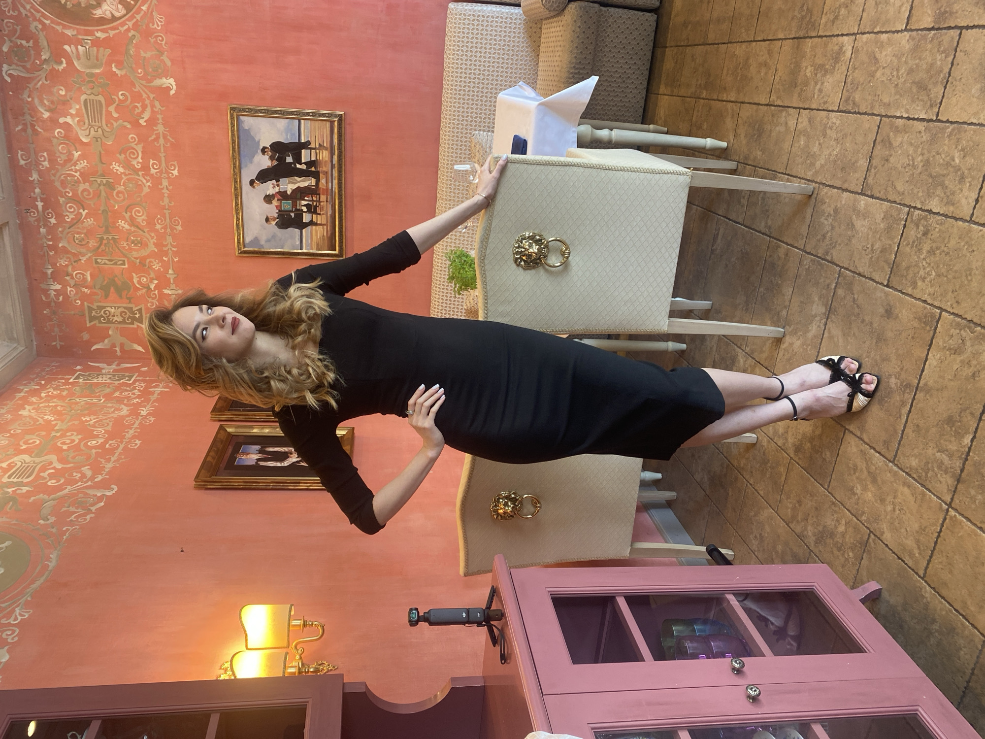 A woman in a black, knee-length dress is posing in what appears to be an upscale indoor setting, possibly a restaurant or dining room. She has long, wavy blonde hair and is looking upwards and to her left. Her right hand rests on her hip, while her left arm extends to lean on the back of a beige upholstered chair positioned to her right. She is wearing black sandals with gold-toned accents.
The room features reddish-orange walls, with intricate gold stencil patterns adorning the upper sections. Two framed paintings hang on the wall behind the woman; the upper one depicts several figures in dark suits, possibly an orchestra or a group of men gathered. The lower painting is less distinct.
The floor is covered with large, light brown rectangular tiles. Two upholstered chairs with decorative gold lion head rings on their backs are visible, along with a table set with a white tablecloth and a glass. To the lower right of the frame, a pink wooden cabinet with glass doors is visible, displaying various items inside. A black gimbal camera on a stand is placed on top of this cabinet. A wall sconce with two illuminated, cream-colored shades is mounted on the wall above the cabinet, casting a warm glow. The overall impression is one of an elegantly decorated interior, likely during daytime given the ambient lighting, though the sconce is also lit.