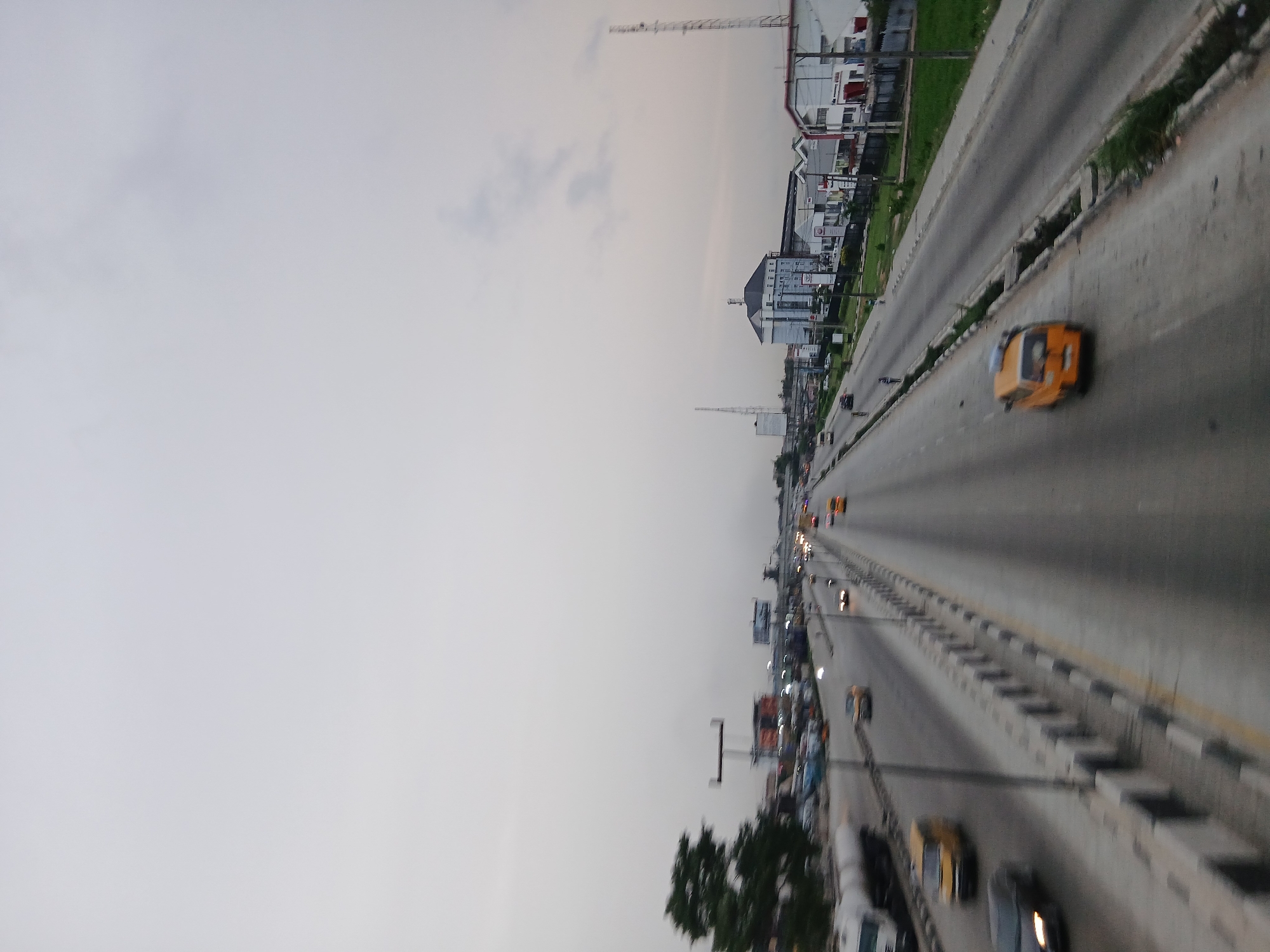 The image depicts a wide highway in an urban area, presumably in Unknown, Unknown. The highway has multiple lanes in each direction, with cars and taxis visible. A median divides the lanes. Buildings are present along the side of the highway. The sky is overcast.