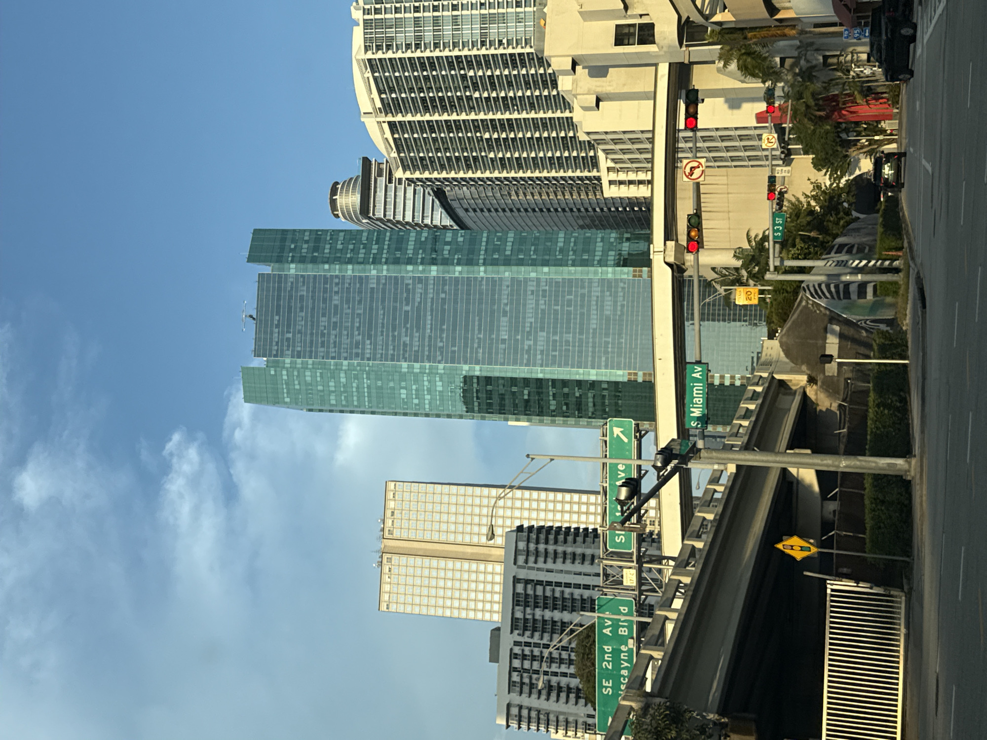 The image shows a street scene in downtown Miami, Florida. Several skyscrapers dominate the skyline. Street signs indicate "SE 2nd Av/ Biscayne Blvd", "S Miami Av", and "S 3 ST". Traffic lights are visible. There is an overpass above the street, and there are vehicles on the road. The sky is blue with scattered clouds.
