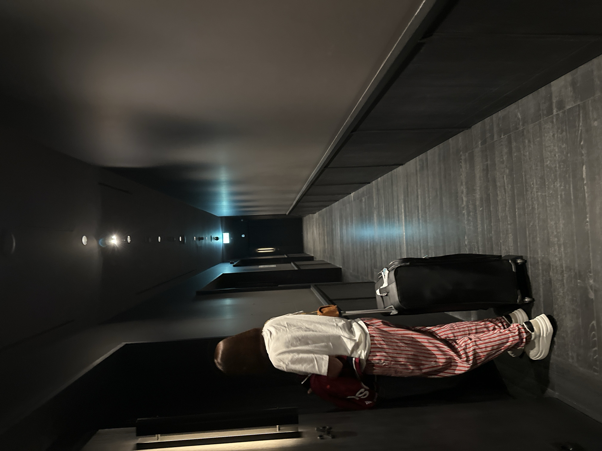 A person in striped pants and a white shirt lies on a dark surface next to a black suitcase. The person also has a red cap and white sneakers. The location appears to be a modern-looking building with dark walls and ceiling, possibly in Tbilisi, Georgia, given the visual aesthetic of the building's architecture and decor. The interior is dimly lit, with a row of lights in the distance.