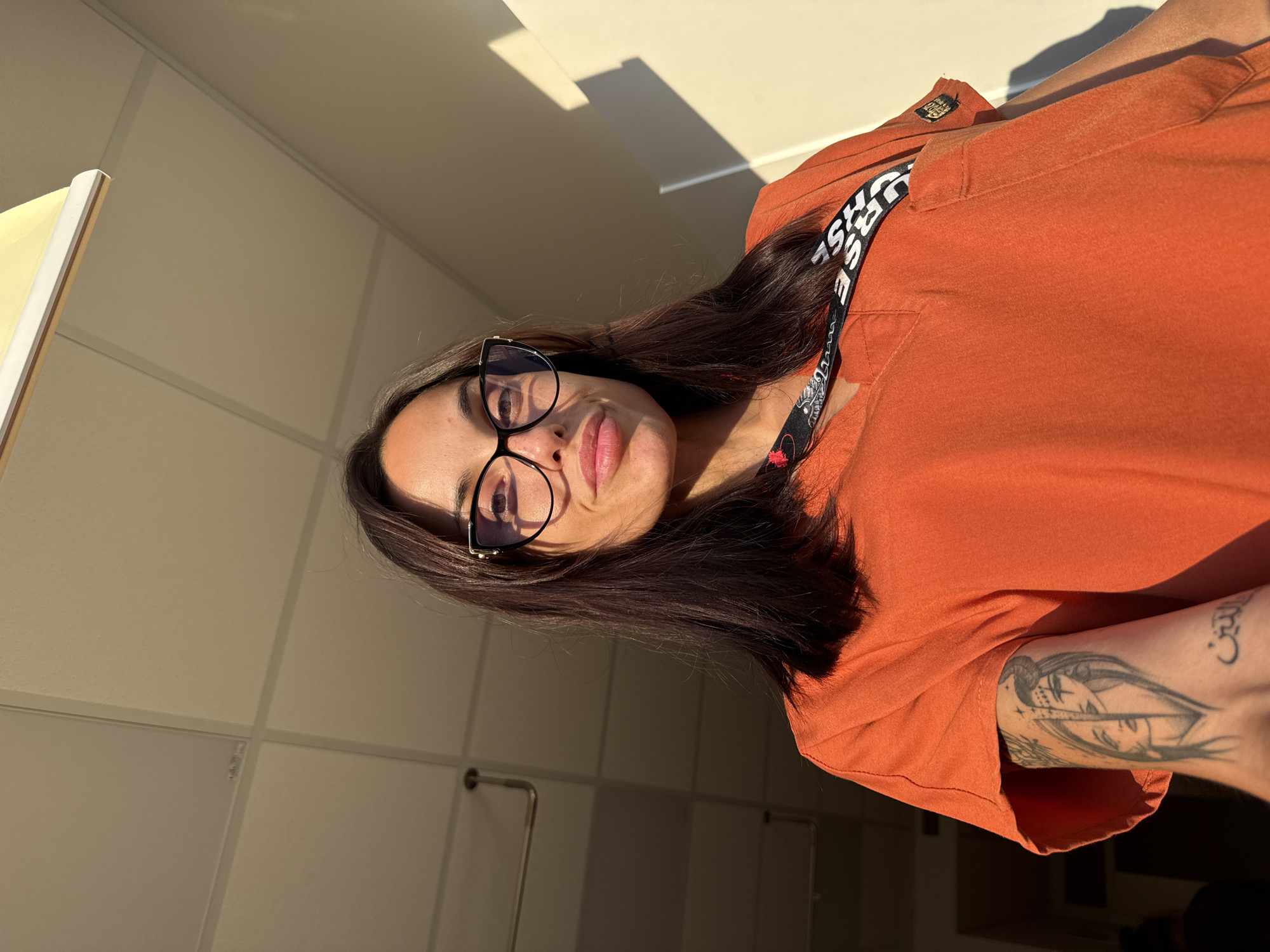 A woman with dark hair and black cat-eye style glasses, featuring gold-colored accents, is depicted in a low-angle indoor photograph. She wears an orange V-neck top, consistent with medical scrubs, and a black lanyard around her neck with partially visible white text reading "URSE". A tattoo on her left upper arm portrays two stylized female faces in a split design, with Arabic script below them, translating to "the religion" or "the faith." Strong, warm, unidirectional light illuminates the scene from the right, creating prominent shadows on the light-colored, possibly beige, wall behind her and highlighting her right side. The ceiling above is a light-colored suspended tile grid with visible metal supports. A linear light fixture is partially visible in the upper right, and a circular pipe or fixture is seen on the upper left. This scene is located in the city of Pleven, Bulgaria.