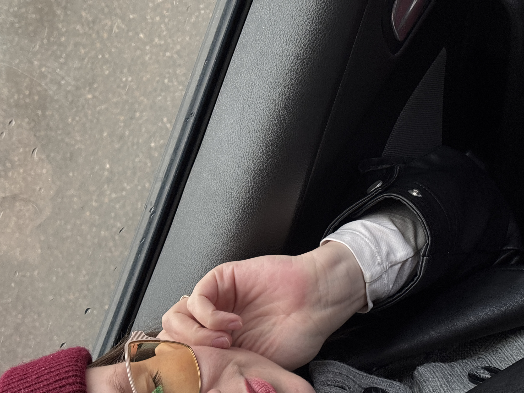 A photograph appears to have been taken from inside a car. The window on the left side of the frame is partially visible, showing a wet, grey street outside, potentially indicating rain. To the right of the window and occupying a significant portion of the frame is the interior of the car, predominantly the dashboard or door panel in dark grey or black. The central focus is on a person's hand, clad in a white long-sleeved shirt, resting on a black leather jacket or bag. Adjacent to the hand is a portion of the person's face, including the nose and mouth, suggesting a relaxed or resting posture. The person is wearing sunglasses with gold frames and a dark red knitted hat or head covering is visible. There is no specific information in the image to pinpoint the location as Chaikovsky, Russia or any other place.