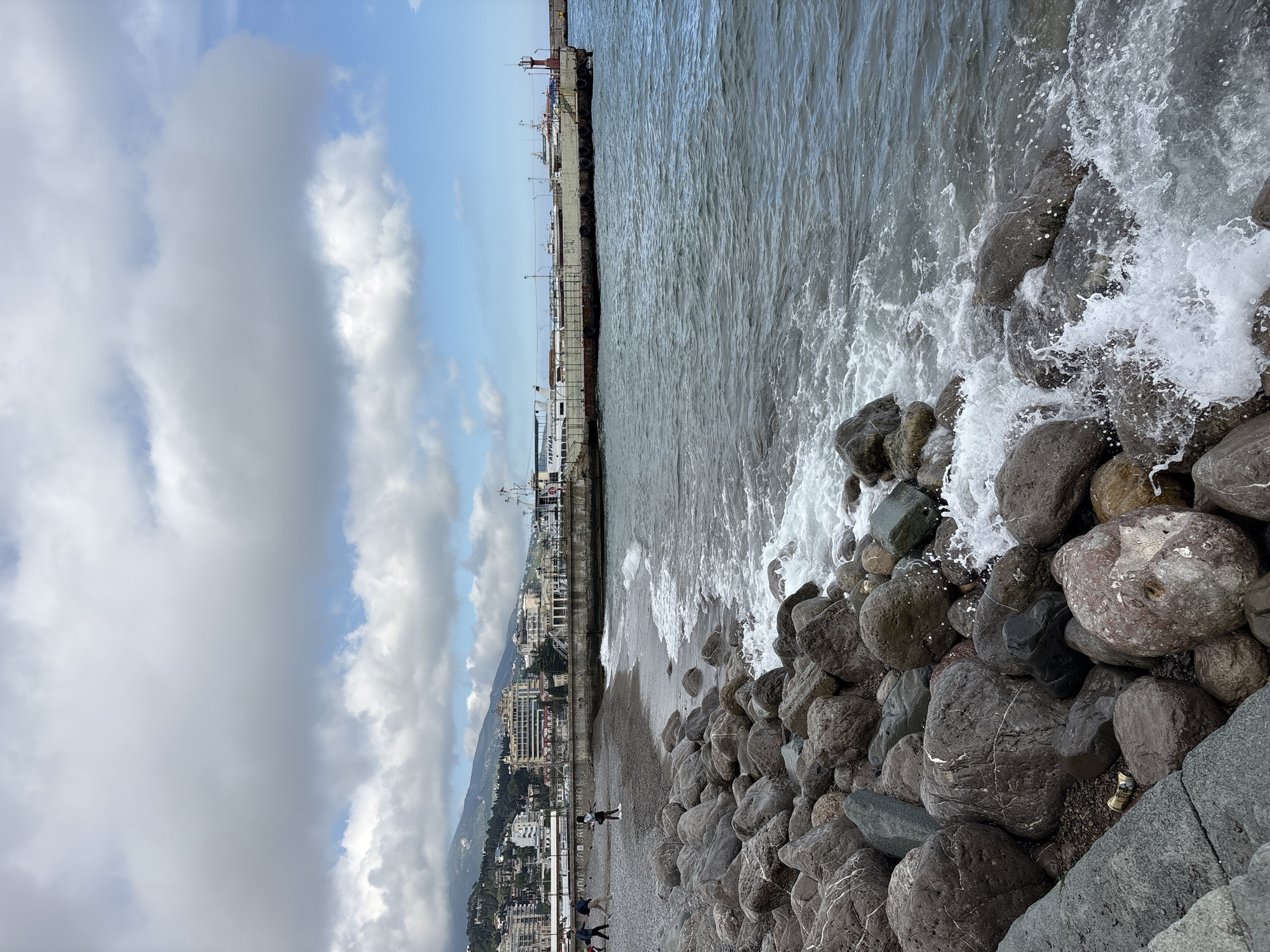 The image shows a coastal scene in город Ялта, Ukraine. In the foreground, large, grey rocks line the shore, where waves crash against them. The shoreline transitions into a narrow beach. Further inland, a densely populated cityscape stretches along the coast, backed by forested mountains. The sky is partially cloudy with patches of blue. A pier extends into the sea with several ships docked. People are visible along the beach.