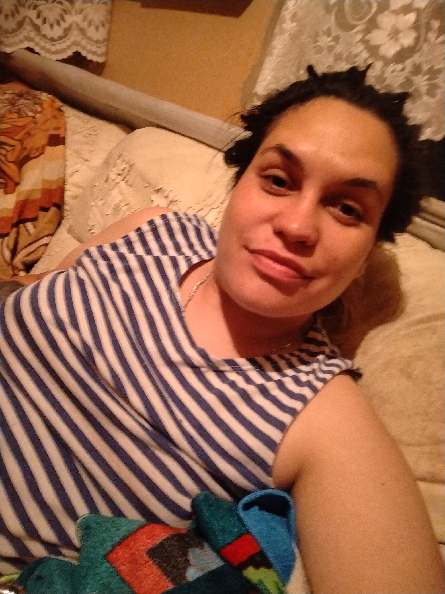 The media file shows a close-up, indoor selfie of a person lying down. The subject is a woman with dark hair pulled up, wearing a blue and white striped tank top and a silver necklace. She has fair skin and appears to be smiling slightly. The background suggests a domestic setting, possibly a bedroom, with a beige textured surface that could be a bed or couch. There are hints of other objects in the background, including what looks like a rolled-up bolster pillow and patterned fabric or blankets. The lighting is warm and appears to be artificial, suggesting it is likely evening or night. The overall mood is relaxed and informal. The image is cropped tightly on the subject, with the background elements being out of focus. No text is visible in the image. The location context is Сосново, Russia.