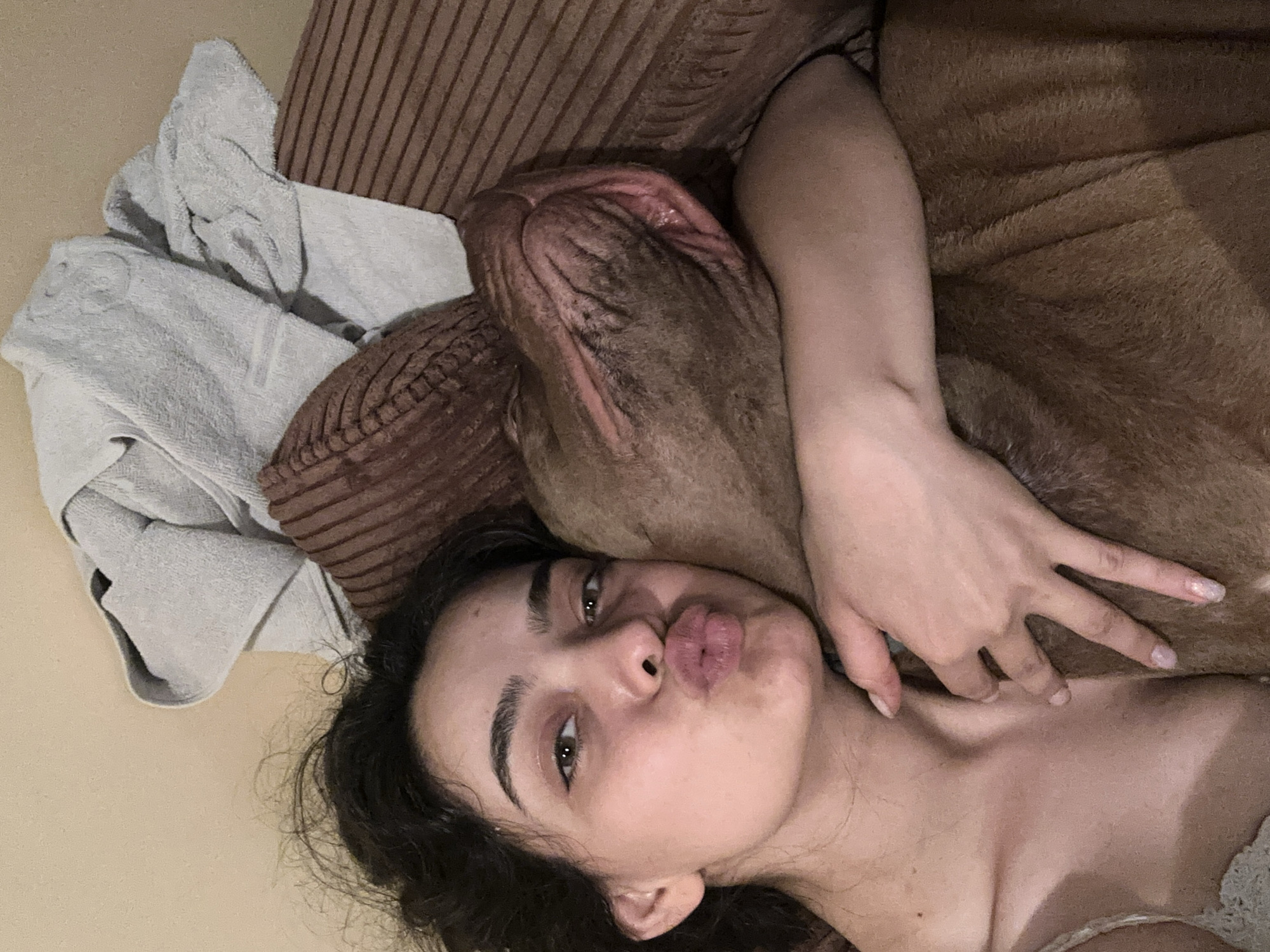 A young woman with dark, curly hair and green eyes is embracing a brown dog. She is making a kissing face towards the camera. The woman is wearing a light-colored lace top. They are lying down on a surface that appears to be a couch with pillows and a folded light-colored towel behind them. The background is a plain, light-colored wall. There is no identifiable information to suggest a location in the city of Malevo, Bulgaria.