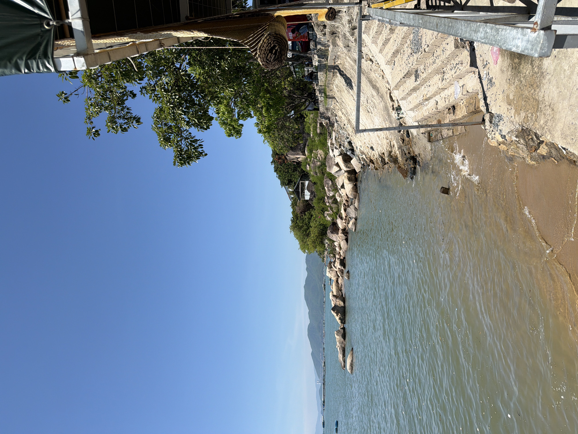 This image depicts a coastal scene in Ninh Ích, Vietnam, on a clear, sunny day. The foreground features a sandy beach meeting the calm, greenish-blue water. To the right, a concrete embankment with rough-hewn steps leads up from the water. This area appears to be part of a waterfront establishment, with a corrugated metal roof and exposed beams visible overhead, and a rolled-up mat hanging from a rope. In the midground, a rocky shoreline juts into the sea, with scattered boulders and vegetation. Beyond the rocks, a cluster of buildings and trees are nestled on a hillside, with more distant mountains forming the horizon. The sky is a vibrant, cloudless blue. A few small boats are visible in the water on the left side of the image, and in the background, there appears to be a structure or settlement on the water. A few individuals are casually seated or standing near the buildings on the hillside.
