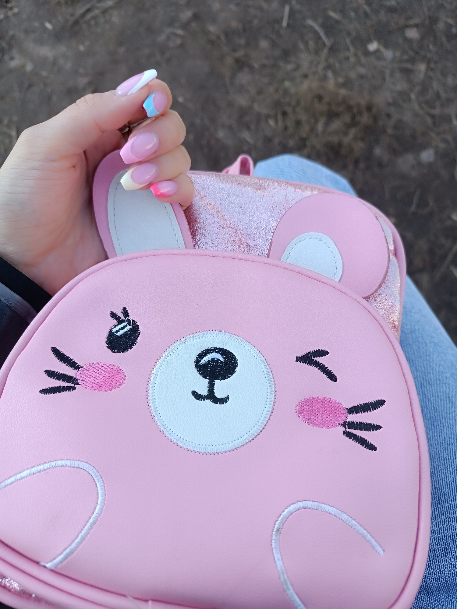 The image shows a person holding a pink, cartoon-like bunny backpack. The backpack has a round, white patch for the face with embroidered details for the eyes, nose, mouth, cheeks, and whiskers. The ears are pink with white inner patches. The person's hand is visible, showcasing colorful manicured nails with different pastel shades on each nail. The person is wearing blue jeans and is outside, with a blurred background of what appears to be ground or a path. The location is unspecified.
