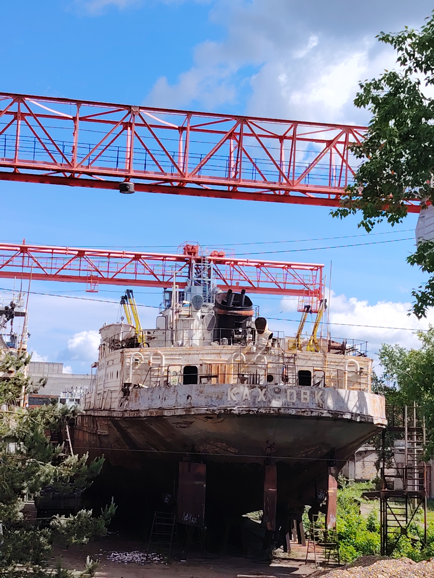 The image depicts a large, rusting ship, named "КАХОВКА" (Kakhovka) in Cyrillic letters, raised on supports in an industrial setting, likely a shipyard. Two red overhead cranes span the area above the ship. The sky is visible between the cranes, with scattered clouds on a bright day. Vegetation surrounds the base of the ship. The location appears to be the city of Chaikovsky, Russia, based on the ship's name.