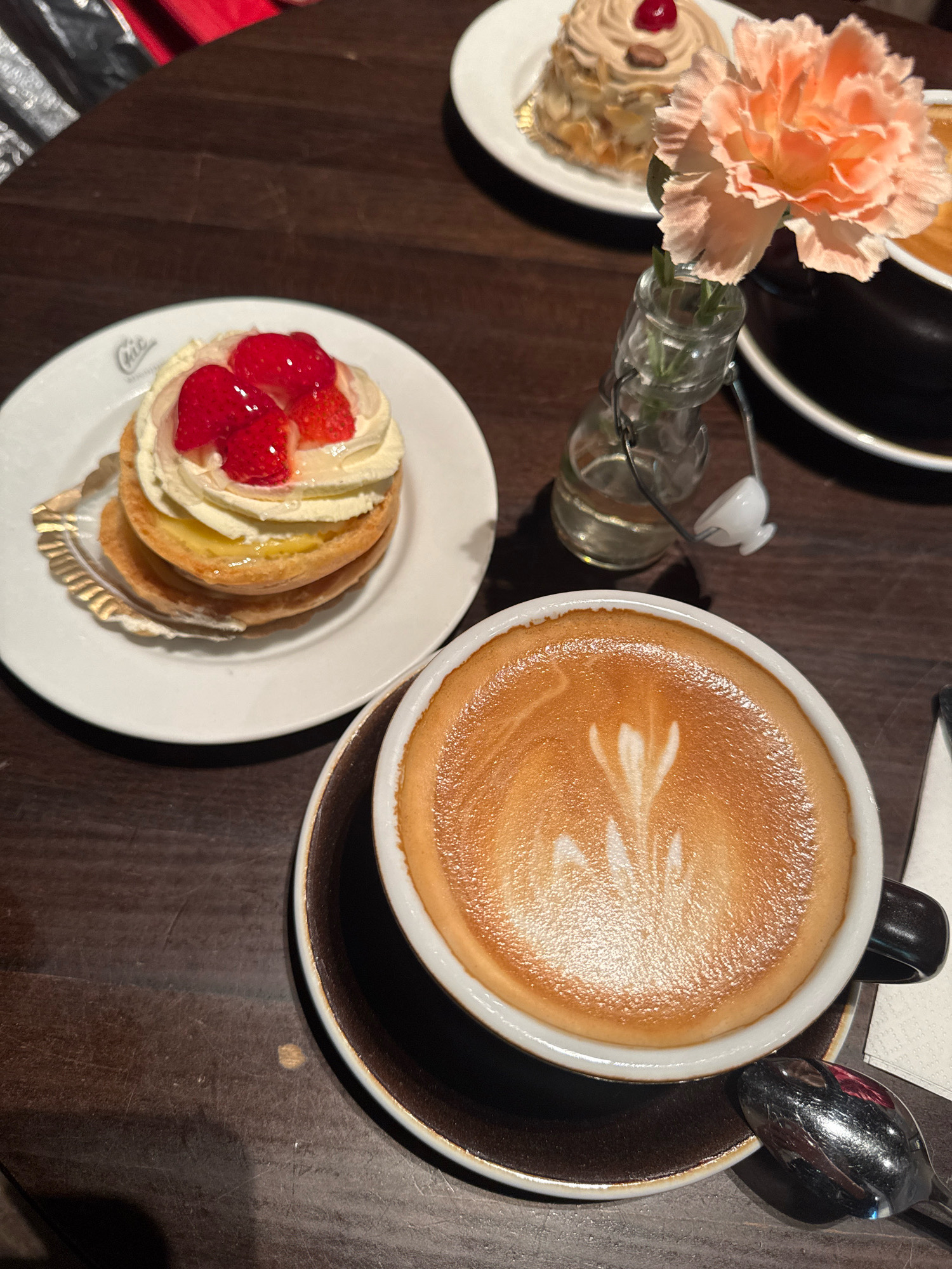 A high-angle shot shows a table with a coffee cup and a pastry, suggesting a cafe setting in Stockholm, Sweden. The coffee cup, filled with a latte adorned with latte art, sits on a dark saucer. To the left, a white plate holds a pastry topped with cream and strawberries. In the background, another pastry with almonds and a cherry is visible. A small glass vase with a single pink carnation adds a decorative touch. The dark wooden table surface is visible throughout the frame, with some wear and tear indicating a well-used space. A silver spoon rests on the saucer to the right of the coffee cup. The lighting is dim, creating shadows that enhance the scene's atmosphere.