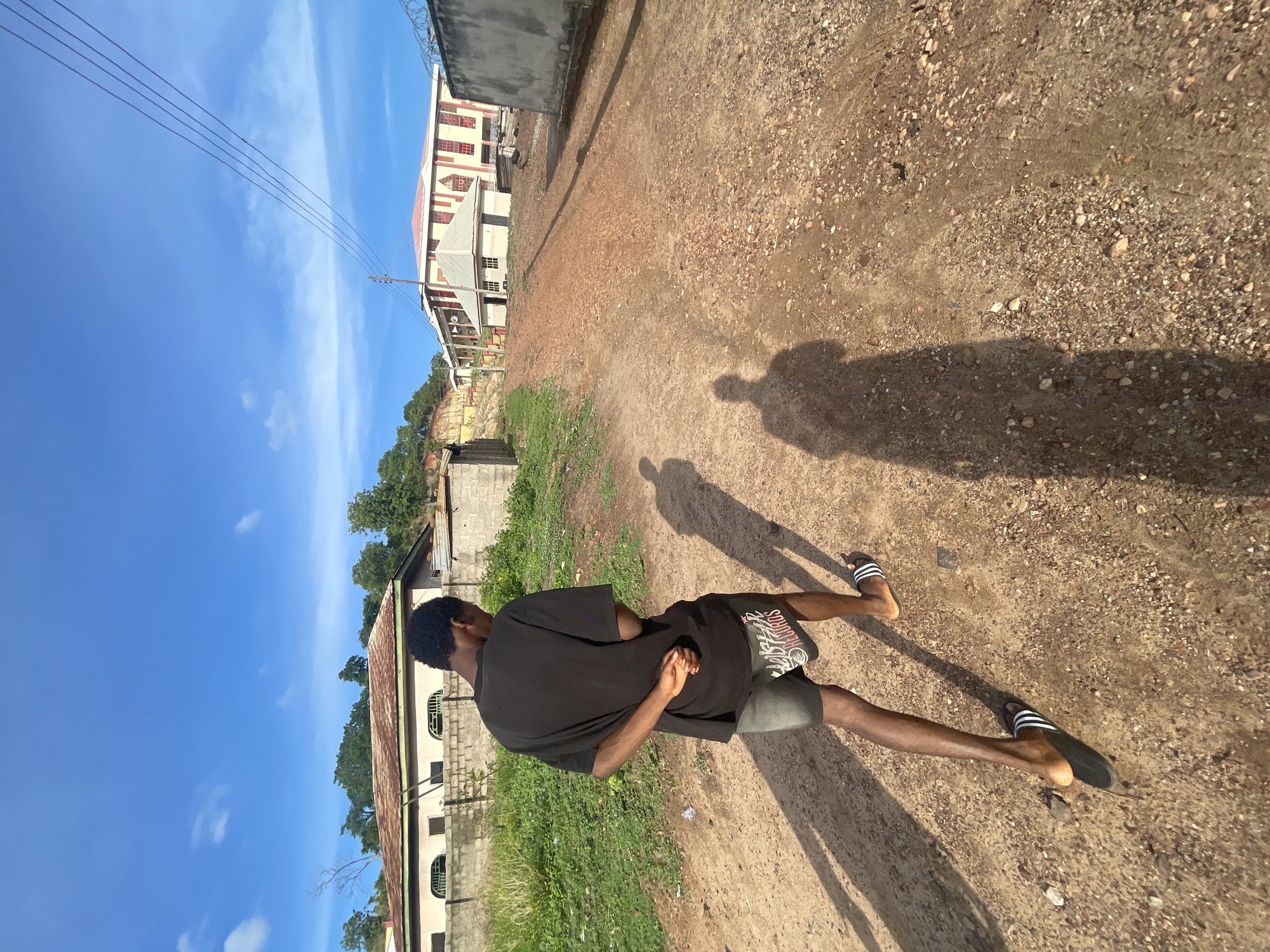 A person stands on a dirt road in Lokoja, Nigeria, with their back to the viewer. They wear a black T-shirt, dark shorts, and black and white striped sandals. The person has short, dark hair. Their hands are clasped behind their back.
The road is dirt with small stones. The person's shadow is cast on the road. To the left, there is a low concrete wall with some vegetation. Behind the wall is a building. To the right, there is another building. Overhead power lines cross the sky. The sky is blue with some clouds.